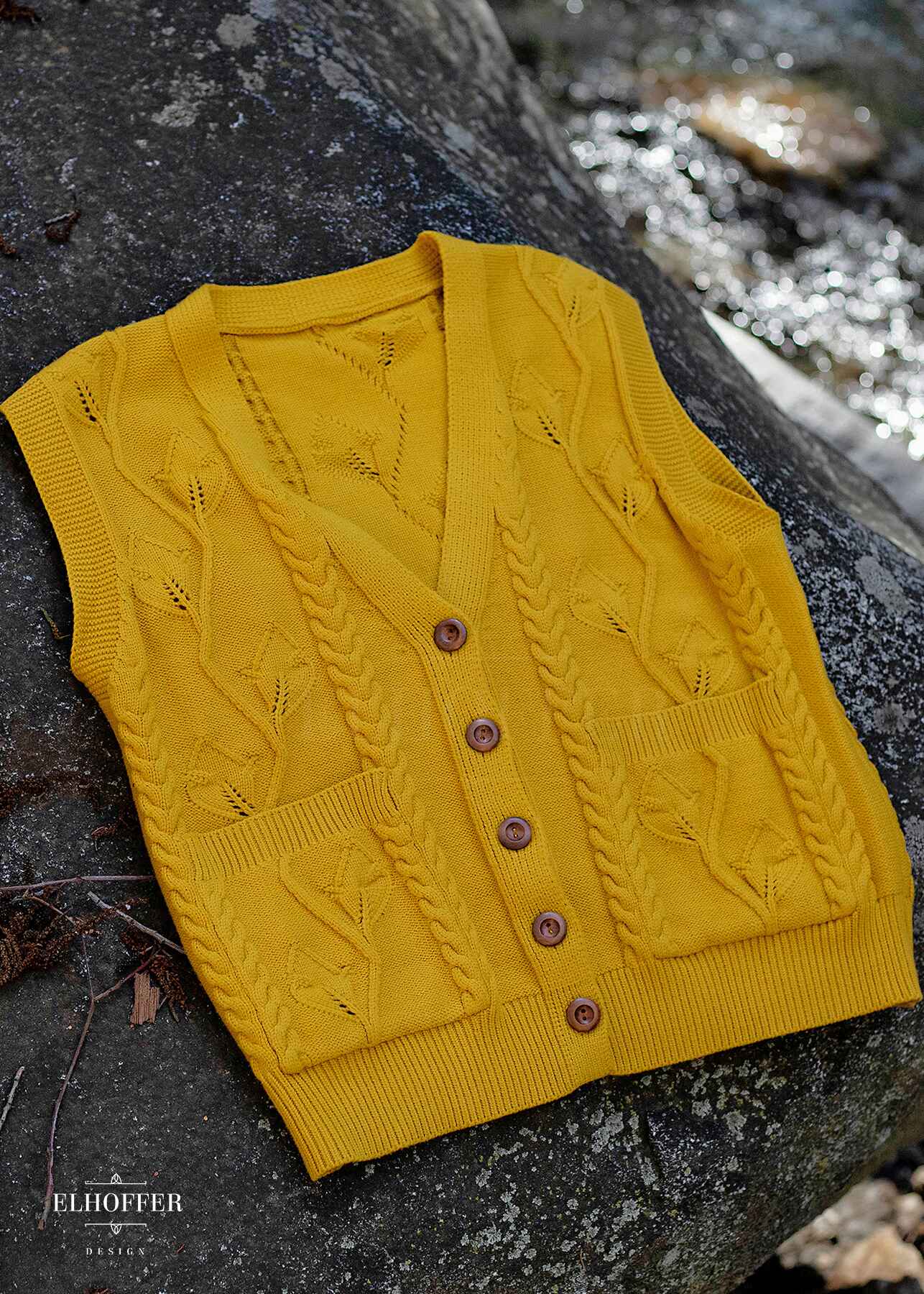 A golden yellow button up knit vest with a leafy vine and cable knit pattern, light brown buttons, and front pockets laying flat on a rock in the woods.