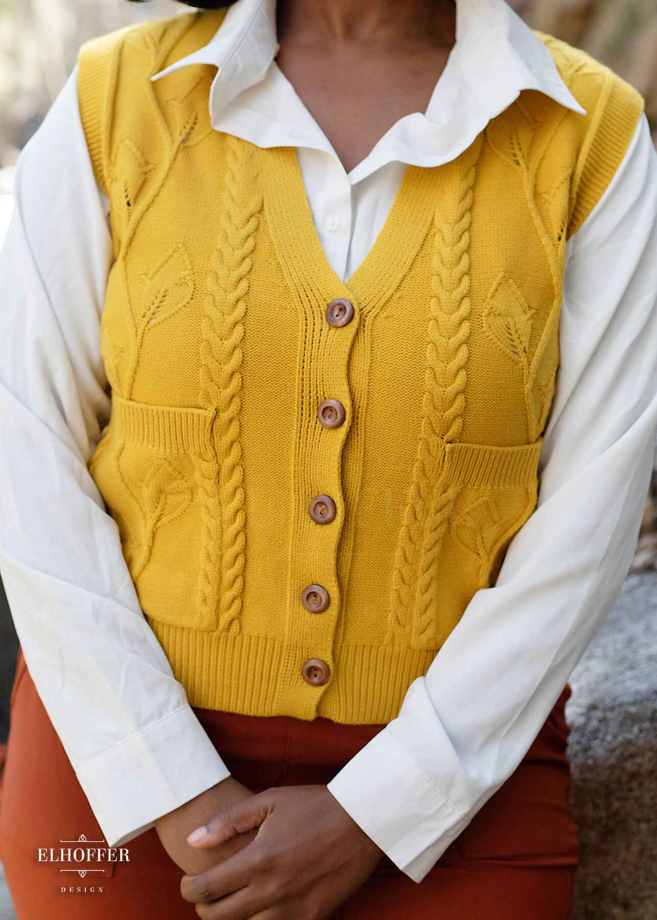 A close up of a golden yellow button up knit vest with a leafy vine and cable knit pattern, light brown buttons, and front pockets.