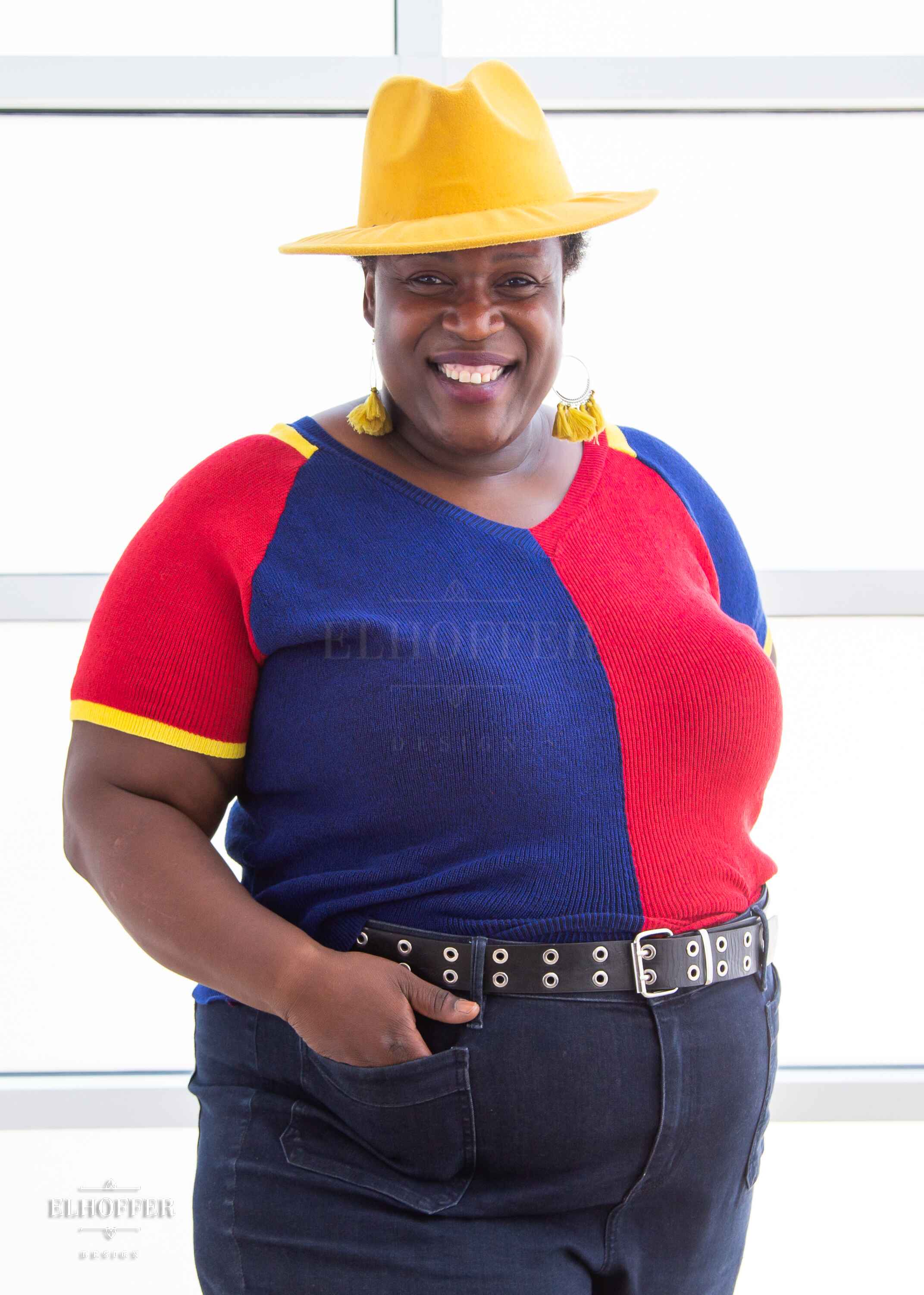 Adalgiza, a medium dark skinned 4xl model with short black super curly hair, is smiling while wearing a short sleeve knit top with alternating blue and red colors.  There is yellow detailing along the top of the shoulder and around the cuff of the sleeve.