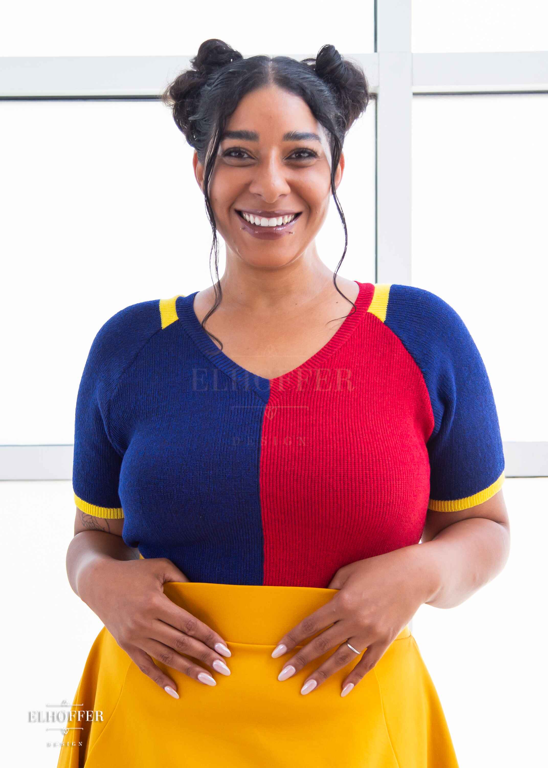 Janae, a light brown skinned L model with dark brown hair in space buns, is smiling while wearing a short sleeve knit top with alternating blue and red colors. There is yellow detailing along the top of the shoulder and around the cuff of the sleeve. She paired the knit top with a golden yellow knee length high low skirt.