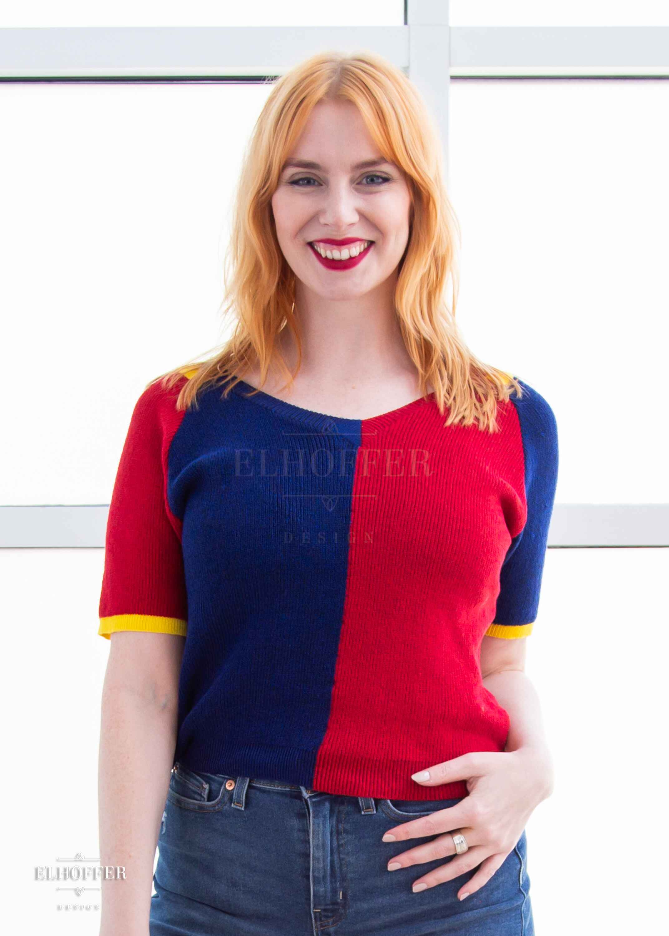 Harley, a fair skinned S model with shoulder length strawberry blonde hair, is smiling while wearing a short sleeve knit top with alternating blue and red colors. There is yellow detailing along the top of the shoulder and around the cuff of the sleeve.