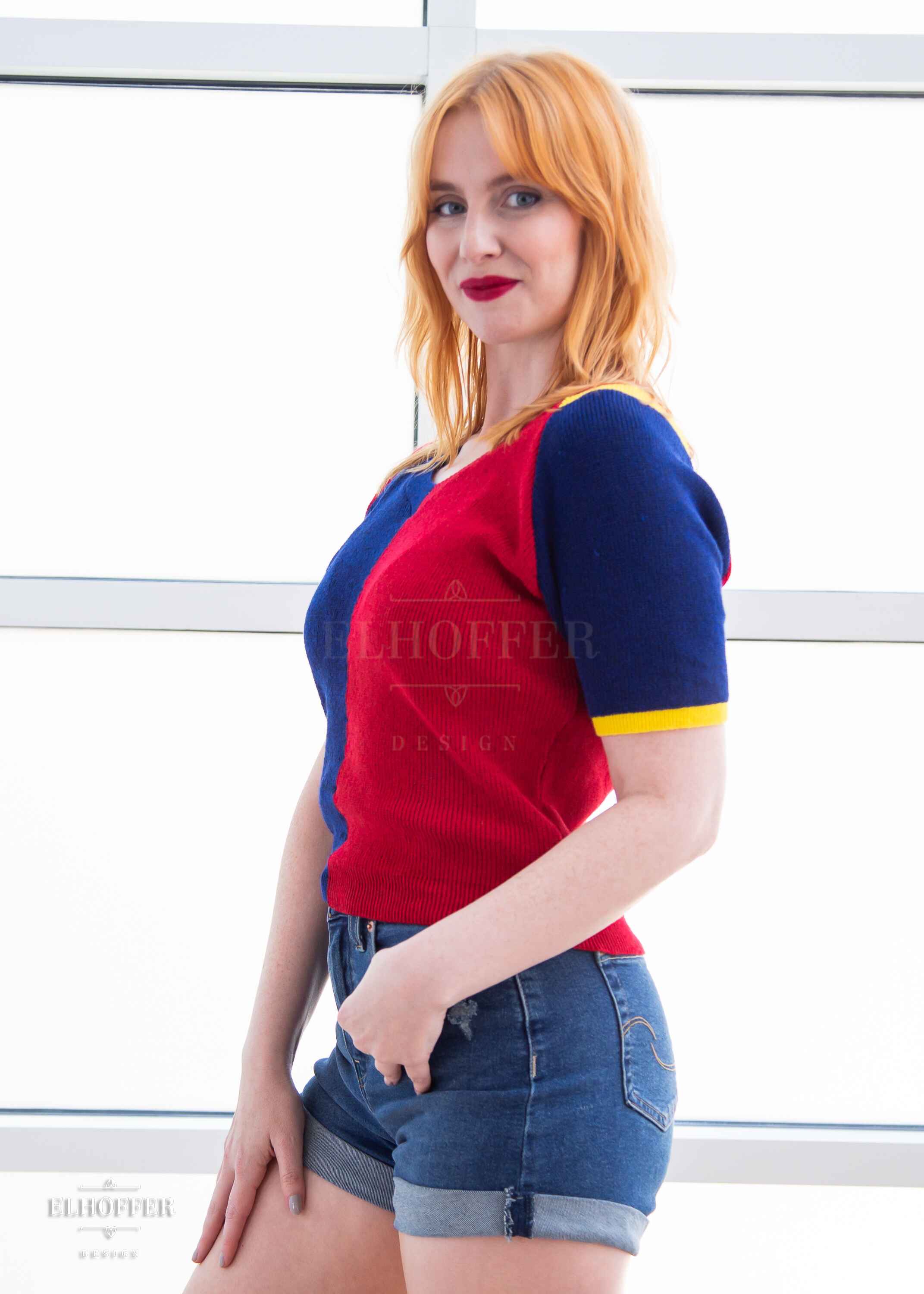 A side view of Harley, a fair skinned S model with shoulder length strawberry blonde hair, wearing a short sleeve knit top with alternating blue and red colors. There is yellow detailing along the top of the shoulder and around the cuff of the sleeve.