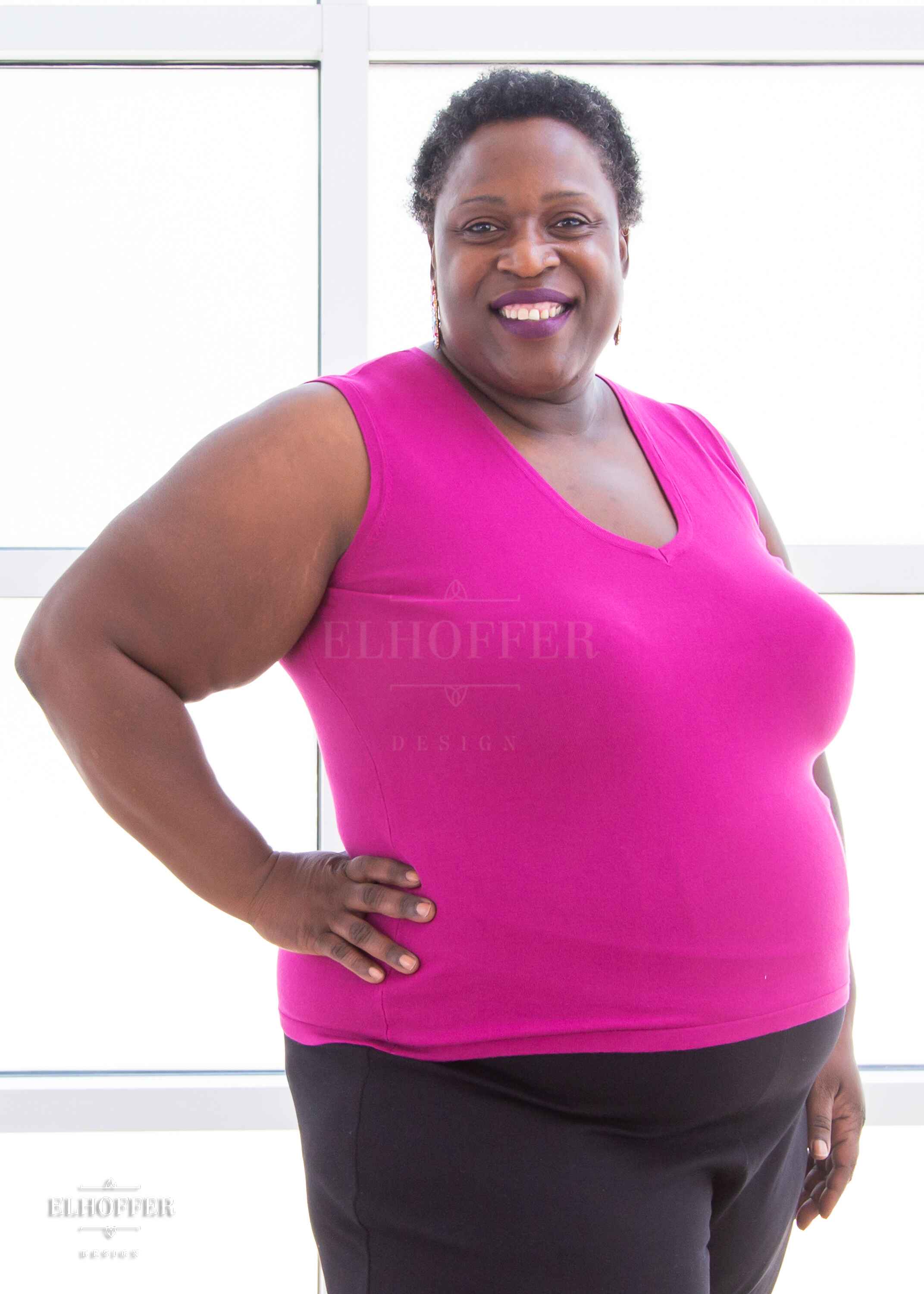 Adalgiza, a medium brown skinned 4xl model with short dark super curly hair, is smiling while wearing a bright pink lightweight knit v neck tank top. The tank top hits about mid hip in length.