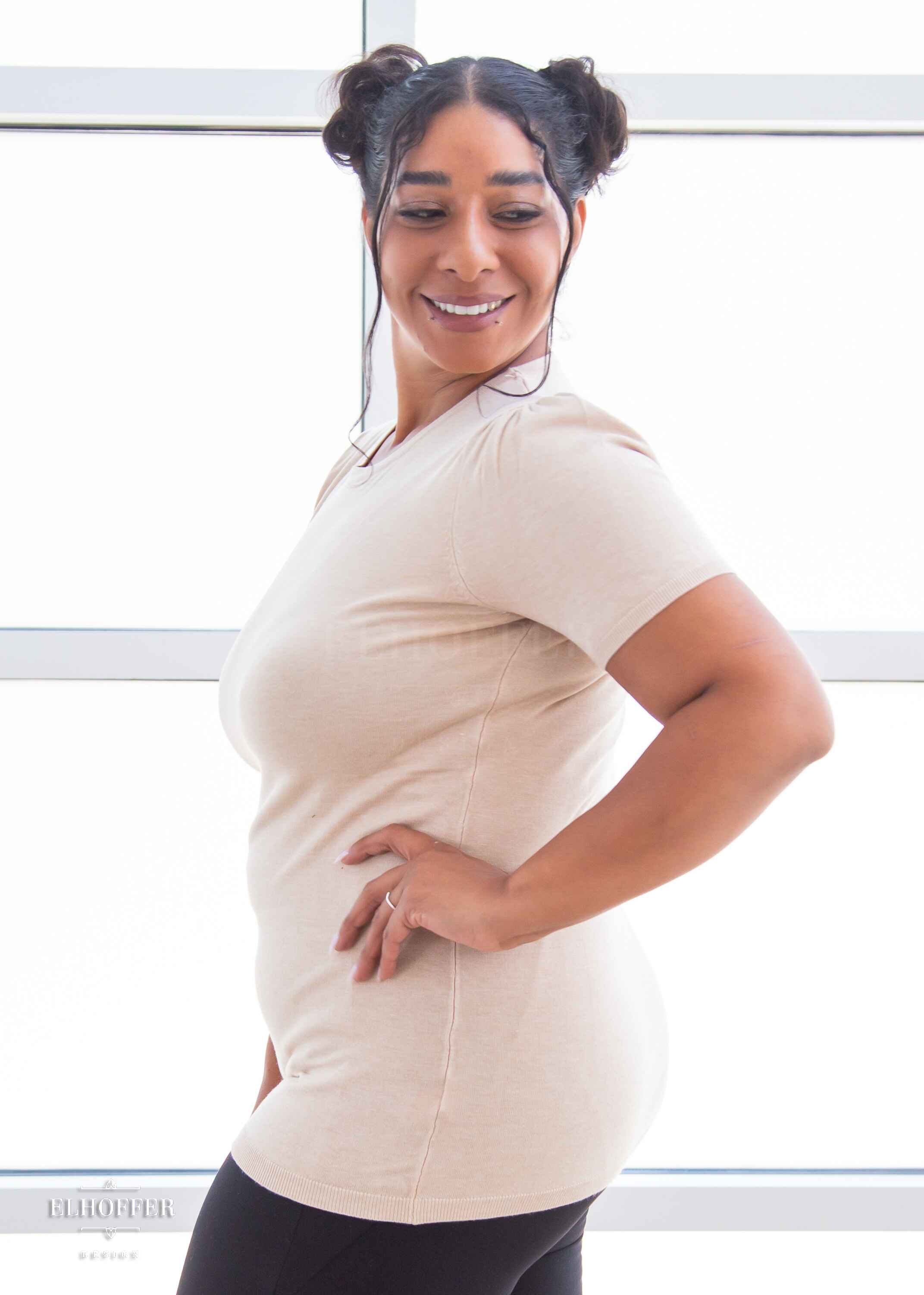 A side view of Janae, a light brown skinned L model with dark hair in space buns, smiling while wearing a short sleeve light weight off white knit top. The top hits about mid hip in length and the sleeves have pleated gathering at the shoulders.