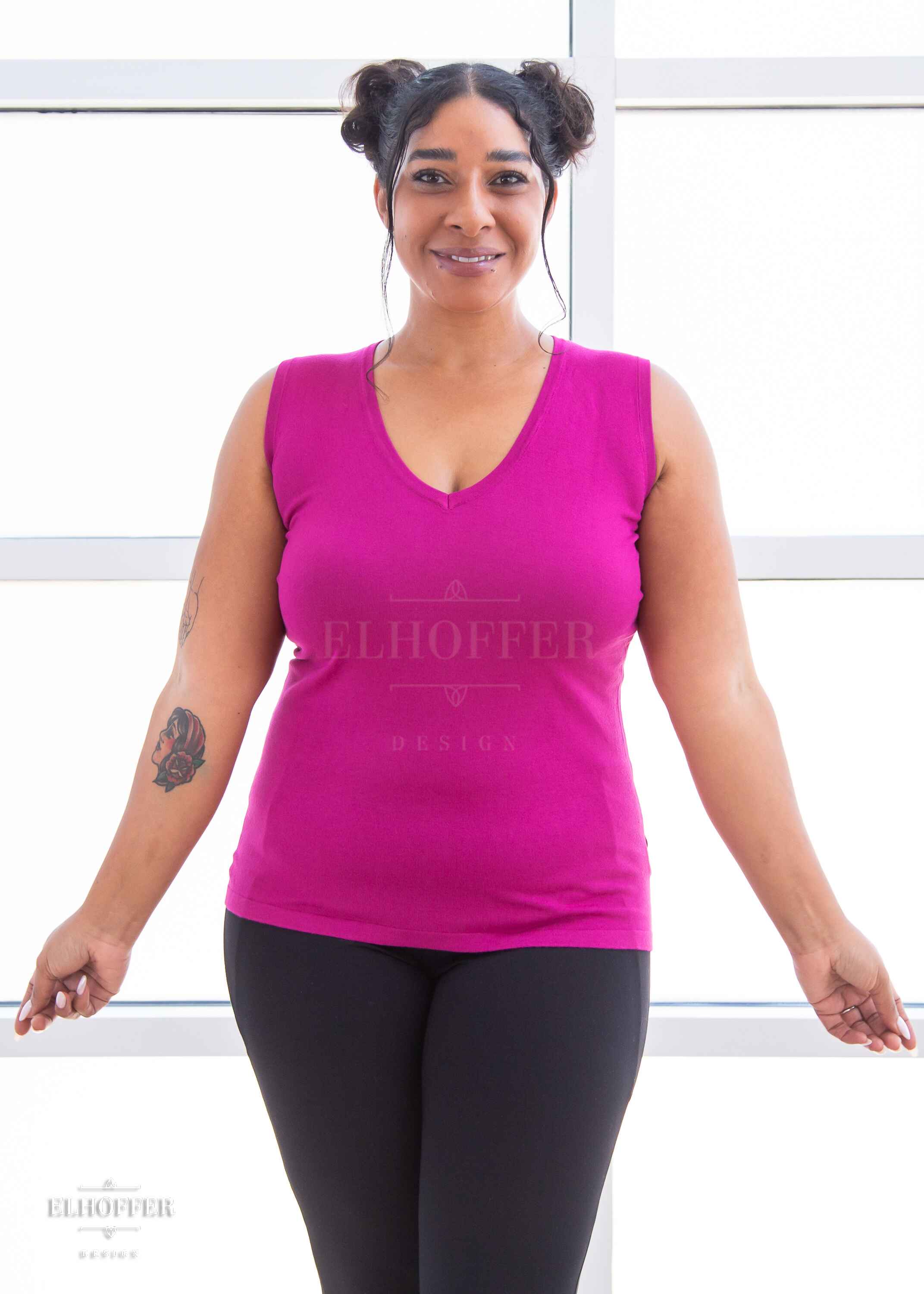 Janae, a light brown skinned L model with dark hair in space buns, is smiling while wearing a bright pink lightweight knit v neck tank top. The tank top hits about mid hip in length.