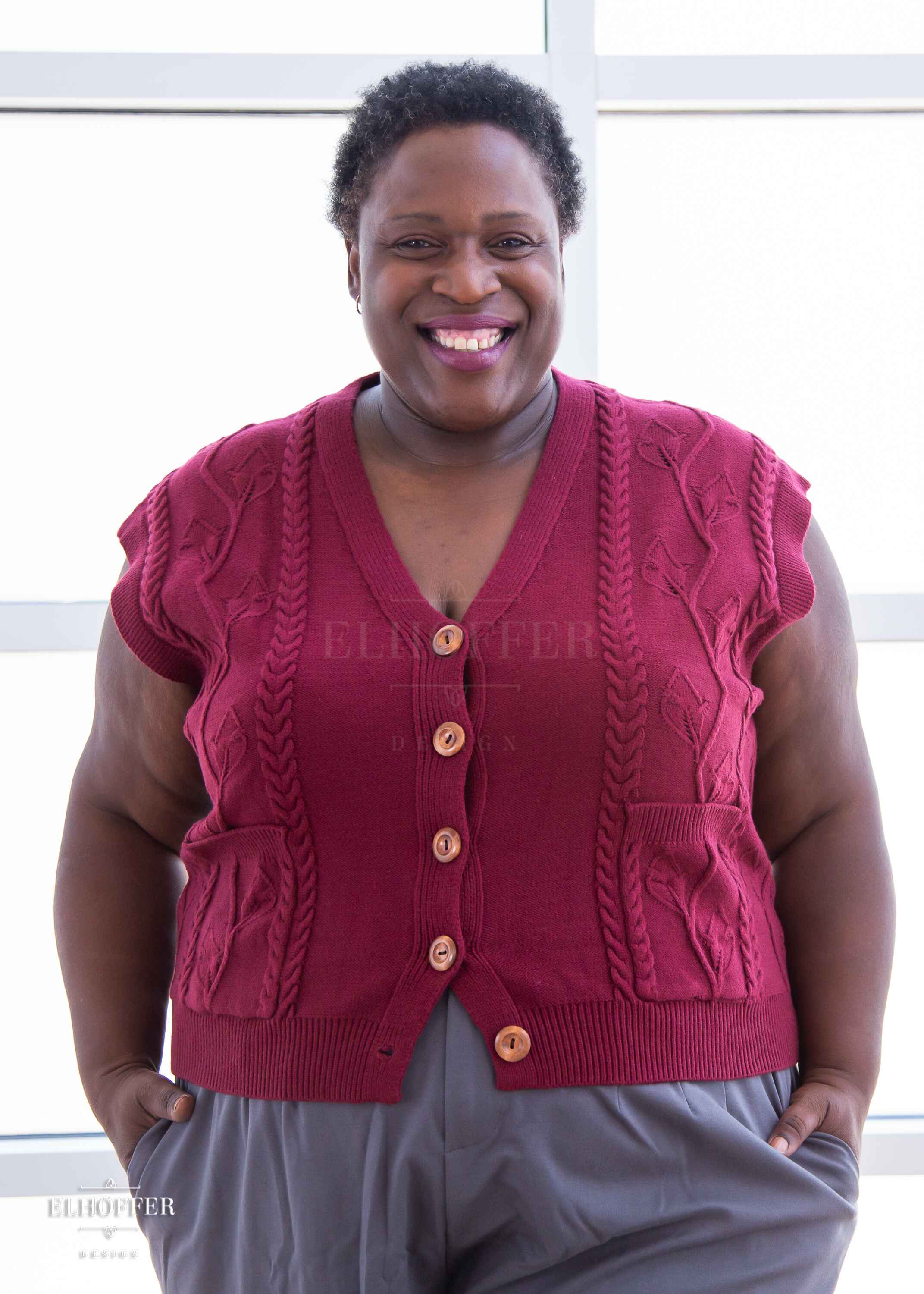 Adalgiza, a medium dark skinned 4xl model with short dark super curly hair, is smiling while wearing the L (3XL-4XL) sample of a cranberry red button up knit vest with a leafy vine and cable knit pattern, light brown buttons, and front pockets. She paired the knit vest with charcoal grey pleated trousers.