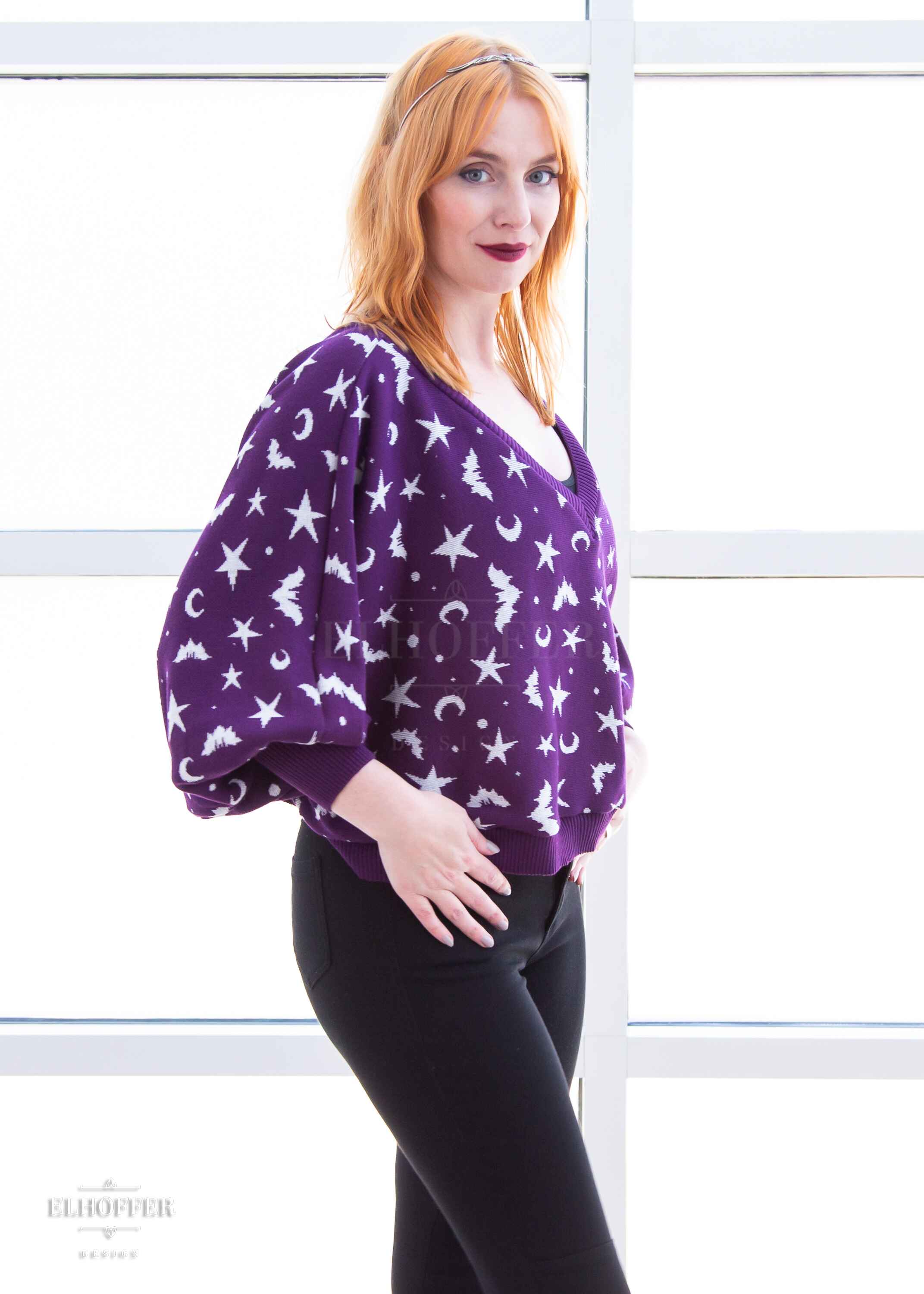 Harley, a fair skinned S model with shoulder length strawberry blonde hair, is wearing an oversized v neck cropped sweater with batwing sleeves that gather at the wrist. The main body of the sweater is purple with a white bat, star, moon, and dot pattern repeated throughout.