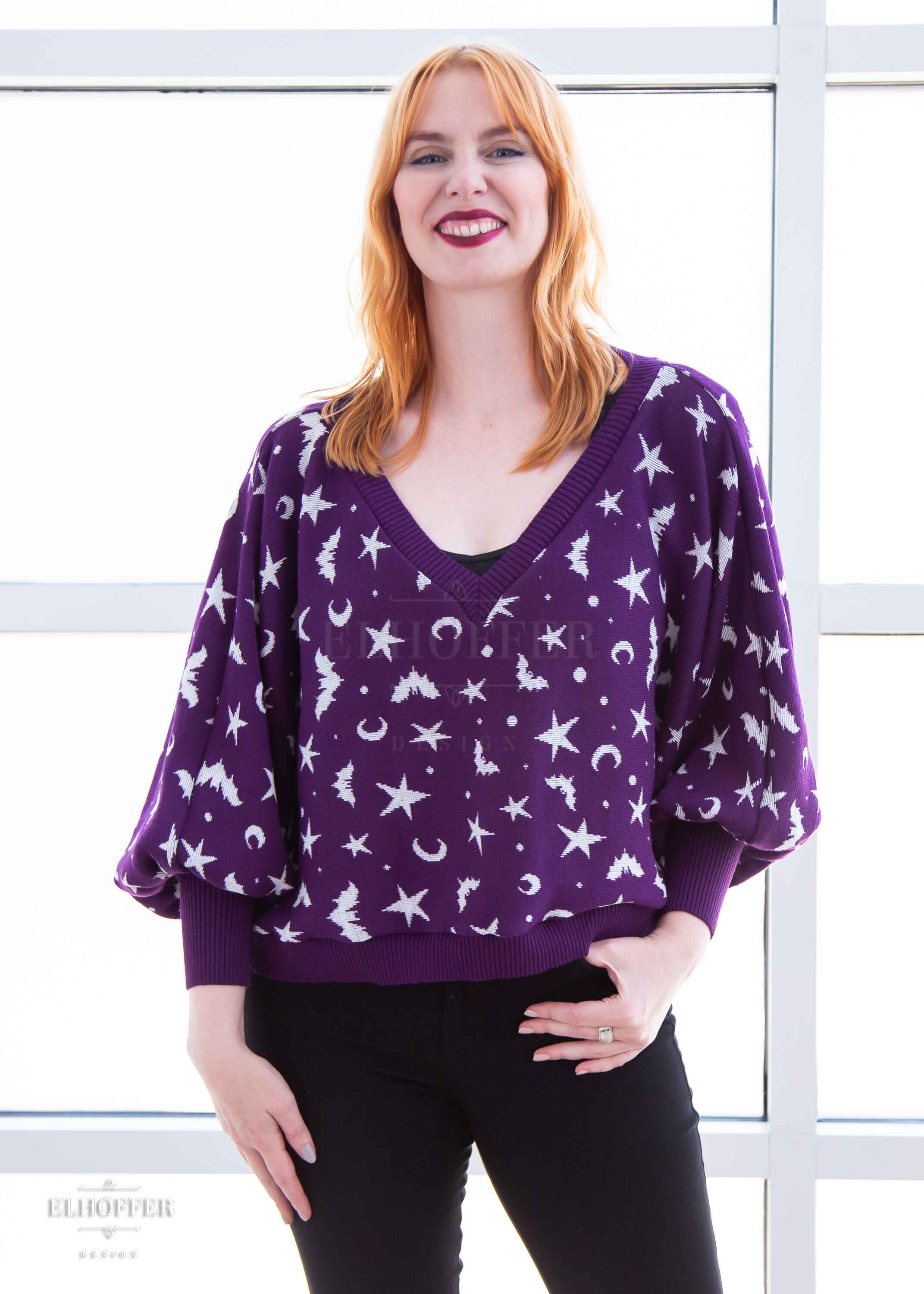 Harley, a fair skinned S model with shoulder length strawberry blonde hair, is smiling while wearing an oversized v neck cropped sweater with batwing sleeves that gather at the wrist. The main body of the sweater is purple with a white bat, star, moon, and dot pattern repeated throughout.