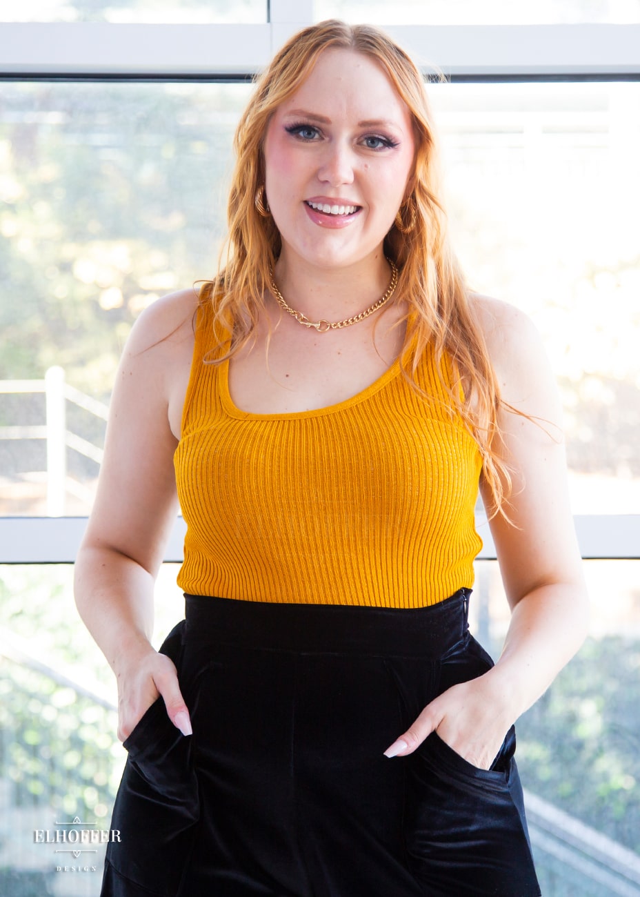 Kelsey, a fair skinned S model with light red hair, is wearing a pullover sleeveless ribbed sweater tank with an asymmetrical neckline in mustard gold tucked in to black velvet pants.