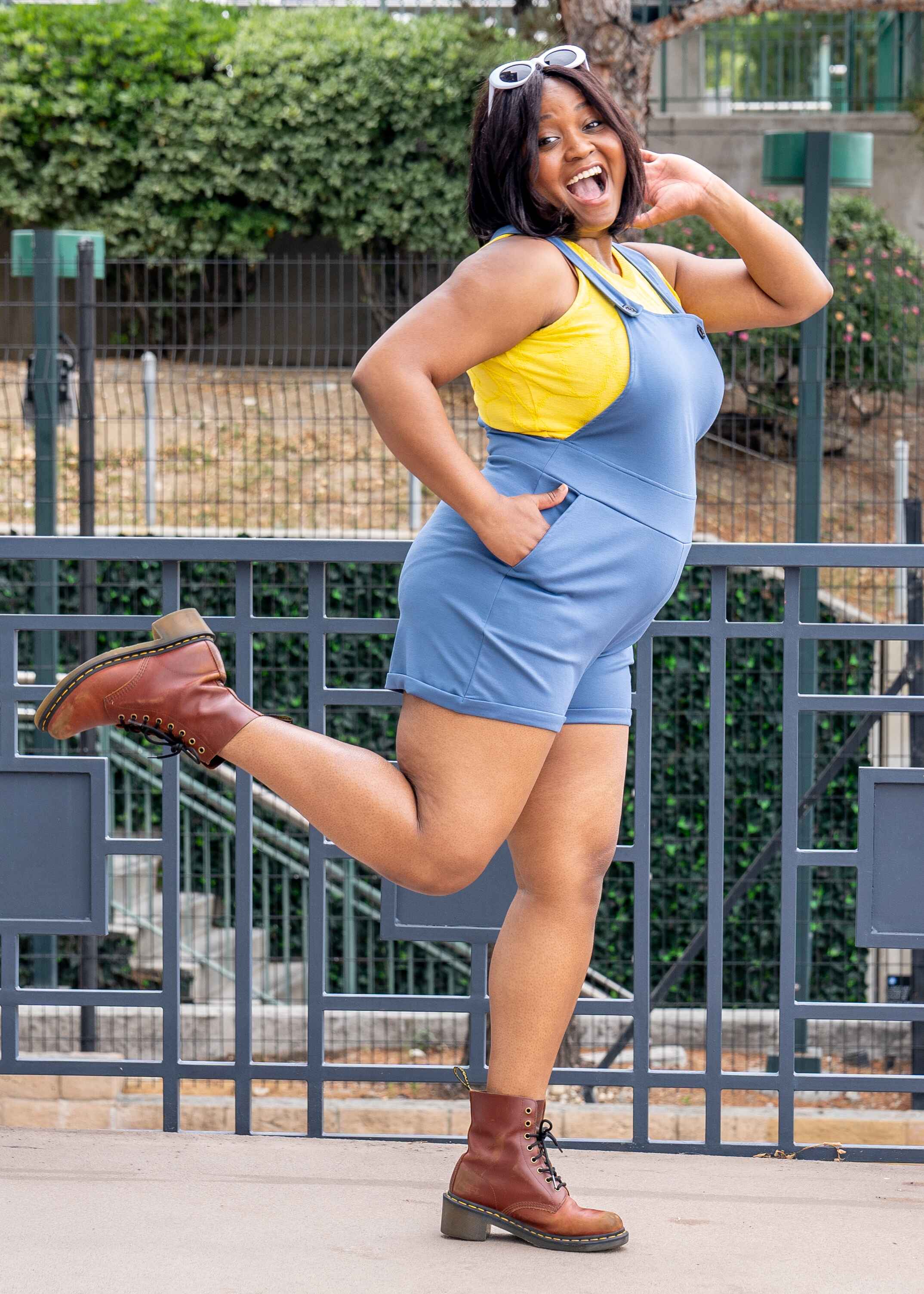 Maydelle, a medium dark skinned XL model with short dark brown hair, is smiling doing the running man while wearing light denim blue short overalls. The overalls have side pockets and multiple buttons on each strap to make it easy to adjust. She paired the overalls with a bright yellow sleeveless knit crop top.