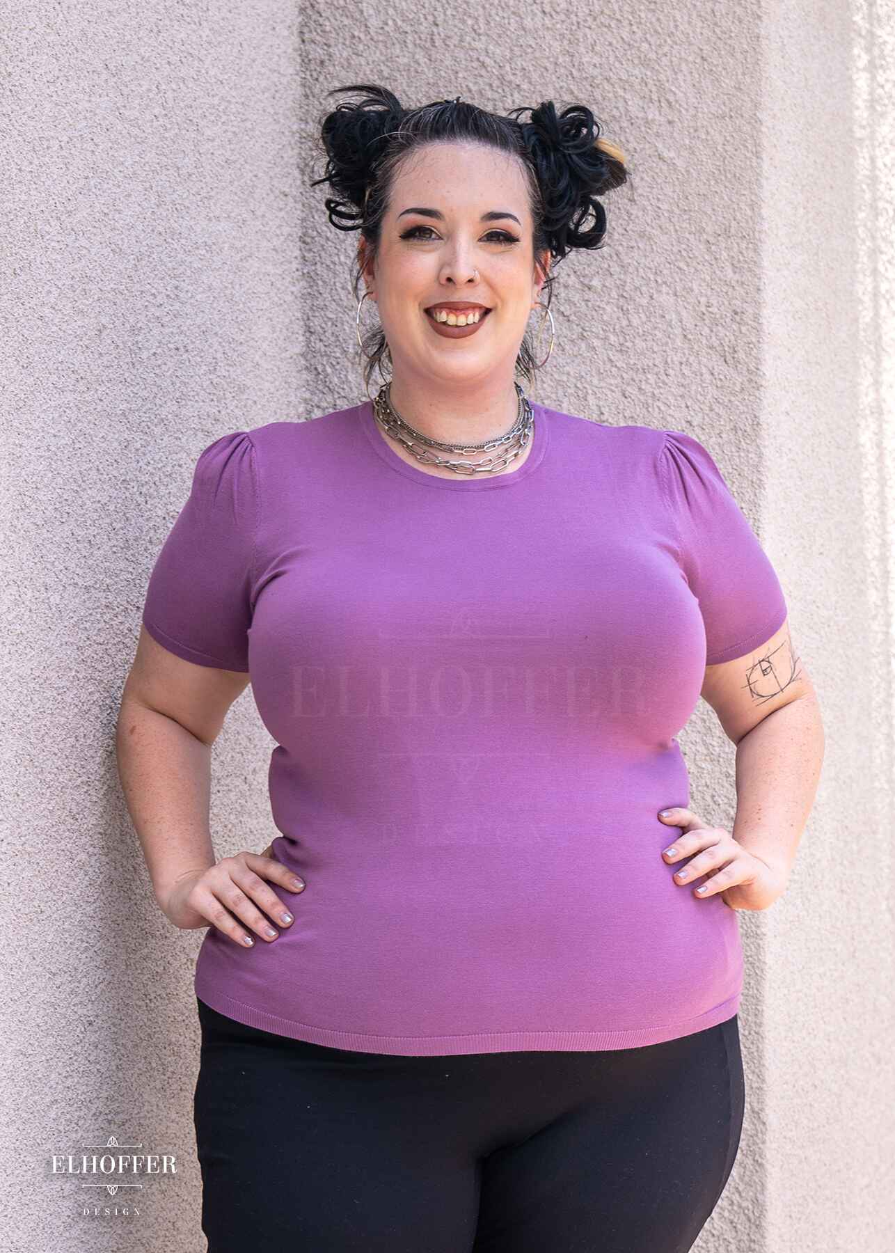 Katie Lynn, a fair skinned 2xl model with dark hair in space buns, is smiling while wearing a short sleeve light weight soft purple knit top. The top hits about mid hip in length and the sleeves have pleated gathering at the shoulders.