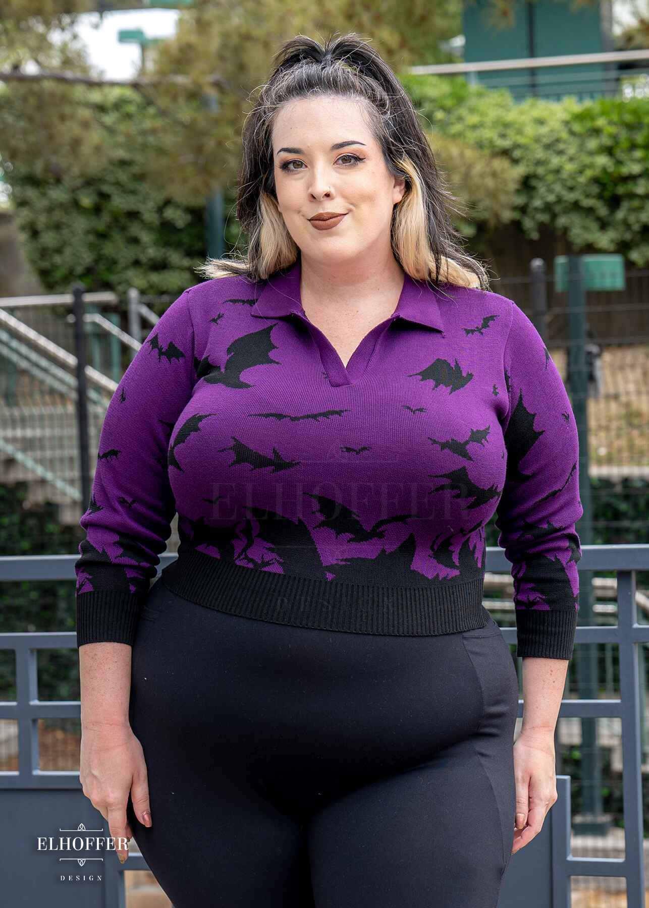 Katie Lynn, a fair skinned 2xl model with shoulder length black and white hair, is wearing the XL sample of a fitted purple cropped 3/4 sleeve knit top with black flying bat design, an open v neck, and collar paired with black leggings. She would normally wear a 2XL for a slightly looser fit.