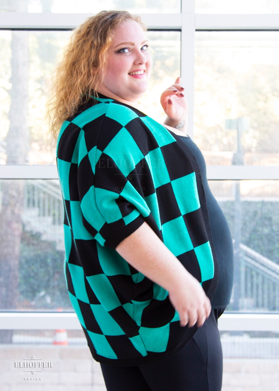 A side view of Bee, a fair skinned 3xl model with shoulder length curly blonde hair, wearing a XL-3XL sample of a shrug with a black and green chessboard pattern. The shrug featured 3/4 sleeves and black ribbing along edges and cuffs.