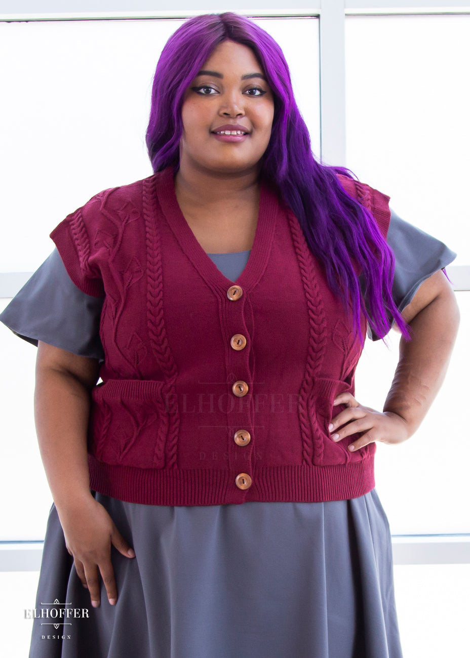 Jade, a light brown skinned 2xl model with long wavy purple hair, is wearing the L (3XL-4XL) sample of a cranberry red button up knit vest with a leafy vine and cable knit pattern, light brown buttons, and front pockets over a charcoal grey knee length dress with short flutter sleeves.