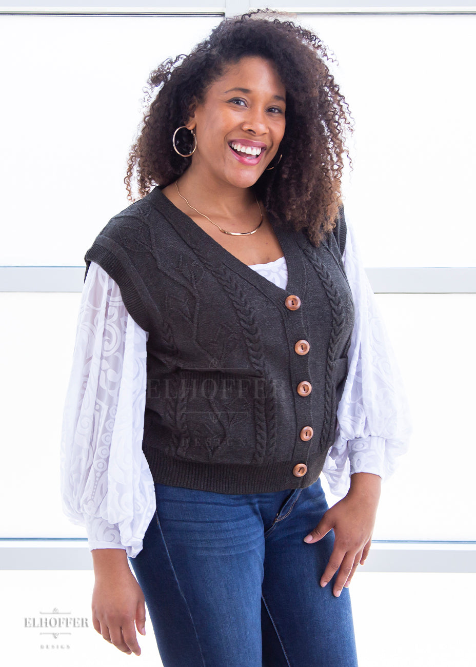 Francesca, a light brown skinned L model with long dark brown tight curly hair, is smiling while wearing the M (L-2XL) sample of a dark grey button up knit vest with a leafy vine and cable knit pattern, light brown buttons, and front pockets over a white long sleeve burnout velvet top with billowing sleeves.