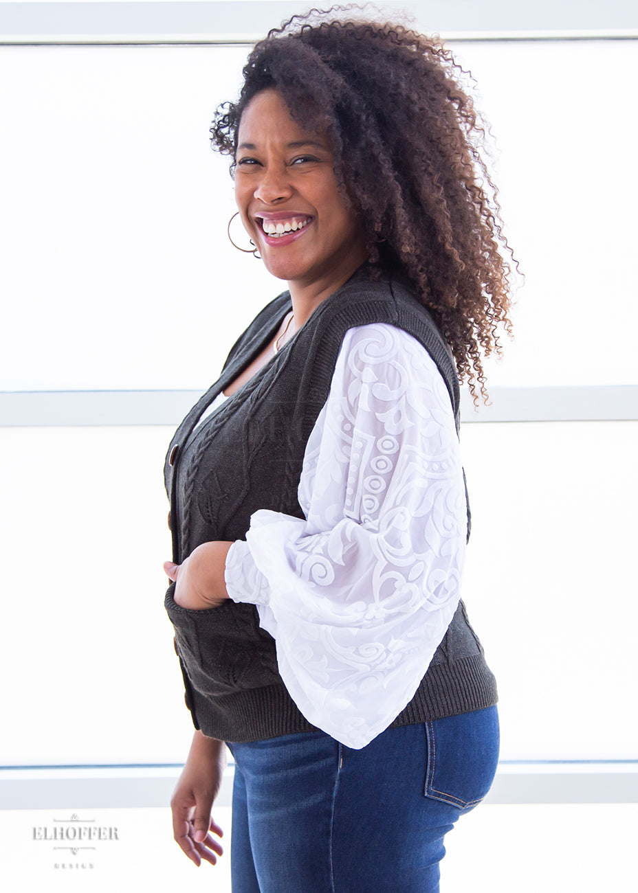Francesca, a light brown skinned L model with long dark brown tight curly hair, has her hand in the pocket of the M (L-2XL) sample of a dark grey button up knit vest she is wearing that has a leafy vine and cable knit pattern and light brown buttons, and she's wearing it over a white long sleeve burnout velvet top with billowing sleeves.