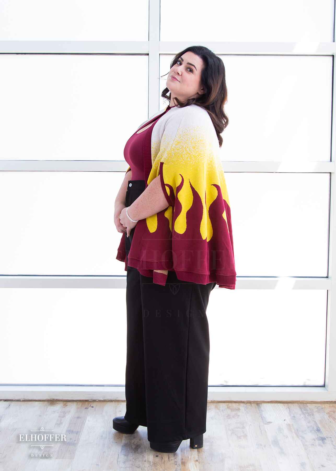 A side view of Stacy, a light skinned XL model with shoulder length wavy dark brown hair, wearing a below hip length knit cape. The cape is a gradient of color from white at the shoulders, to yellow, to a red fire design at the bottom, it also has a button closer at the neck, and armholes in the side seam. She paired the cape with a burgundy scoop neck crop top and black pleated trousers.