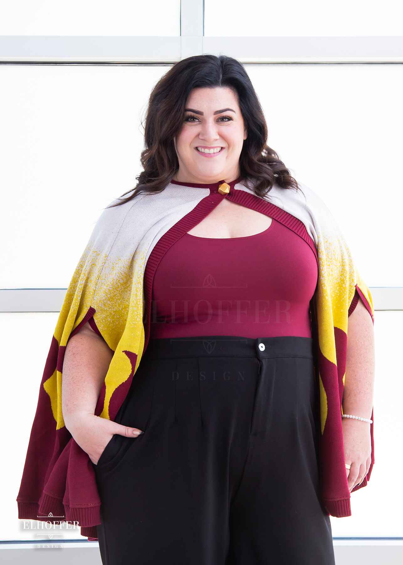 Stacy, a light skinned XL model with shoulder length wavy dark brown hair, is wearing a below hip length knit cape. The cape is a gradient of color from white at the shoulders, to yellow, to a red fire design at the bottom, it also has a button closer at the neck, and armholes in the side seam. She paired the cape with a burgundy scoop neck crop top and black pleated trousers.