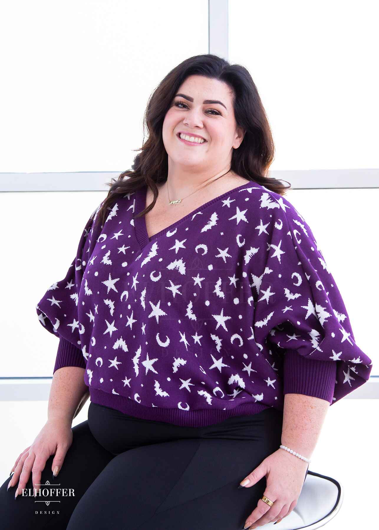 Stacy, a light skinned XL model with shoulder length wavy dark brown hair, is smiling while wearing an oversized v neck cropped sweater with batwing sleeves that gather at the wrist.  The main body of the sweater is purple with a white bat, star, moon, and dot pattern repeated throughout.