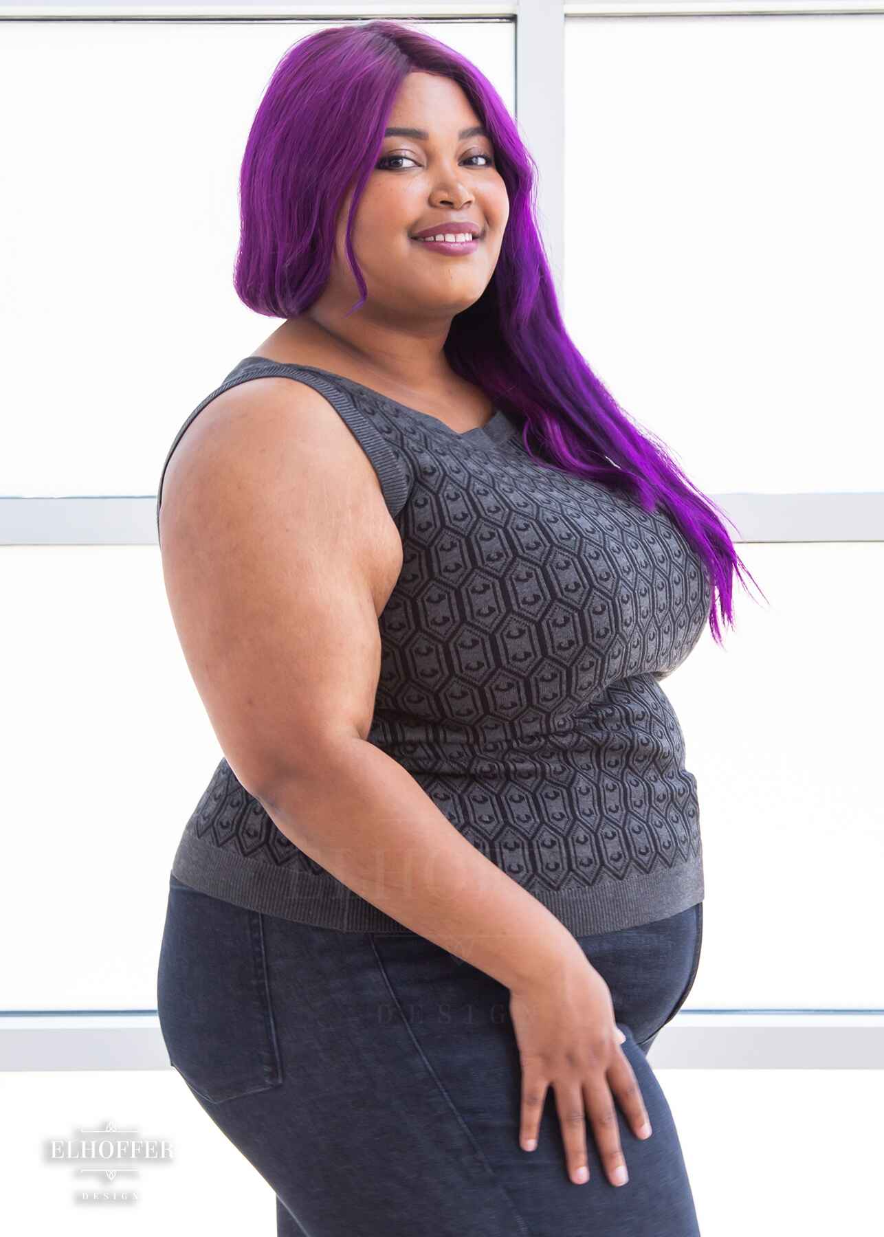 Jade, a light brown skinned 2xl model with long wavy purple hair, is smiling while wearing a knit sleeveless top with a boatneck and an armor plated design
