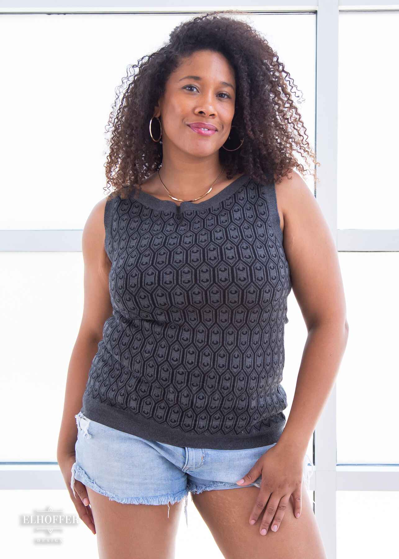 Francesca, a light brown skinned L model with long dark brown tight curly hair, is wearing a knit sleeveless top with a boatneck and an armor plated design.