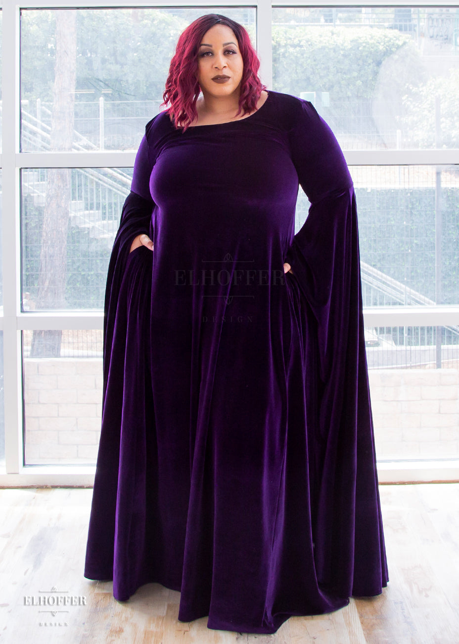 Dawn (a medium dark skinned size 3X model with burgundy hair) wears the purple velvet floor length dress with her hands in the pockets. It has a boatneck neckline and extra long flowing sleeves that fall to ankle length.