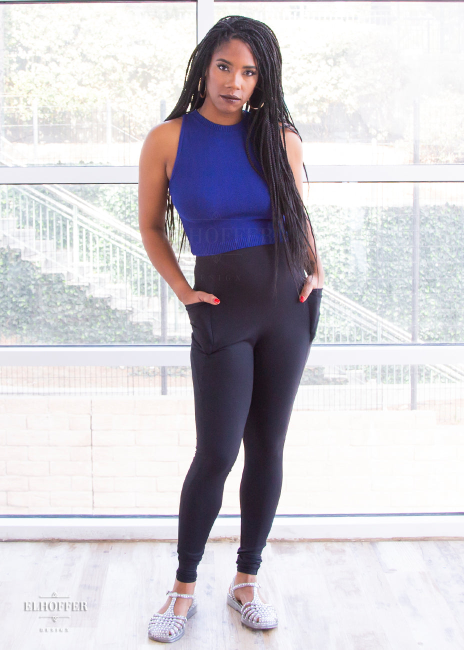 Krystina, a size small medium dark skinned model with long braids, is wearing a pair of high waisted black leggings with a seamless front and side seam pockets in black.