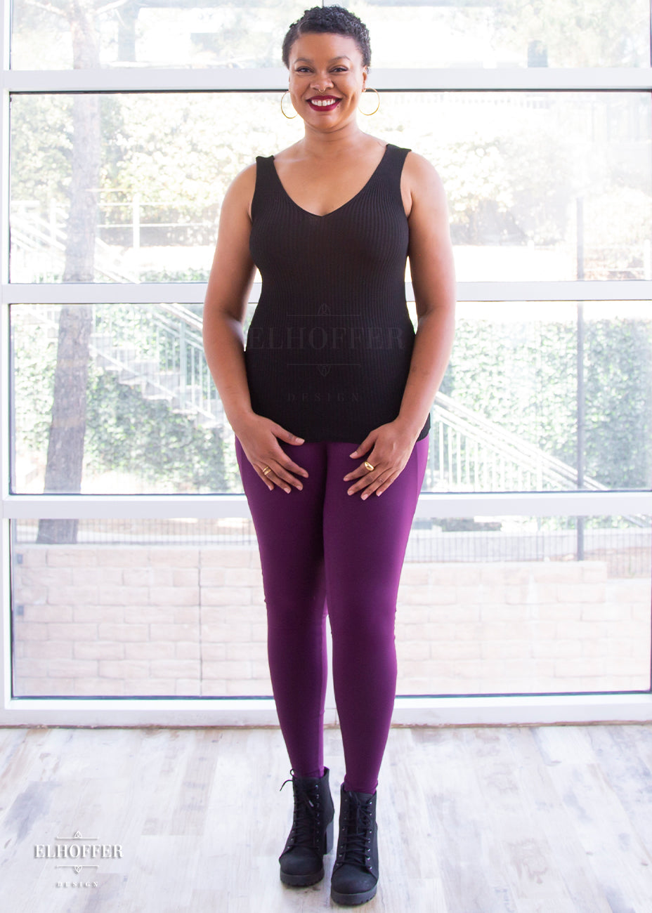 Desiree, a size large medium dark skinned model with very short dark hair, is wearing a pair of high waisted leggings with a seamless front and side seam pockets in plum.