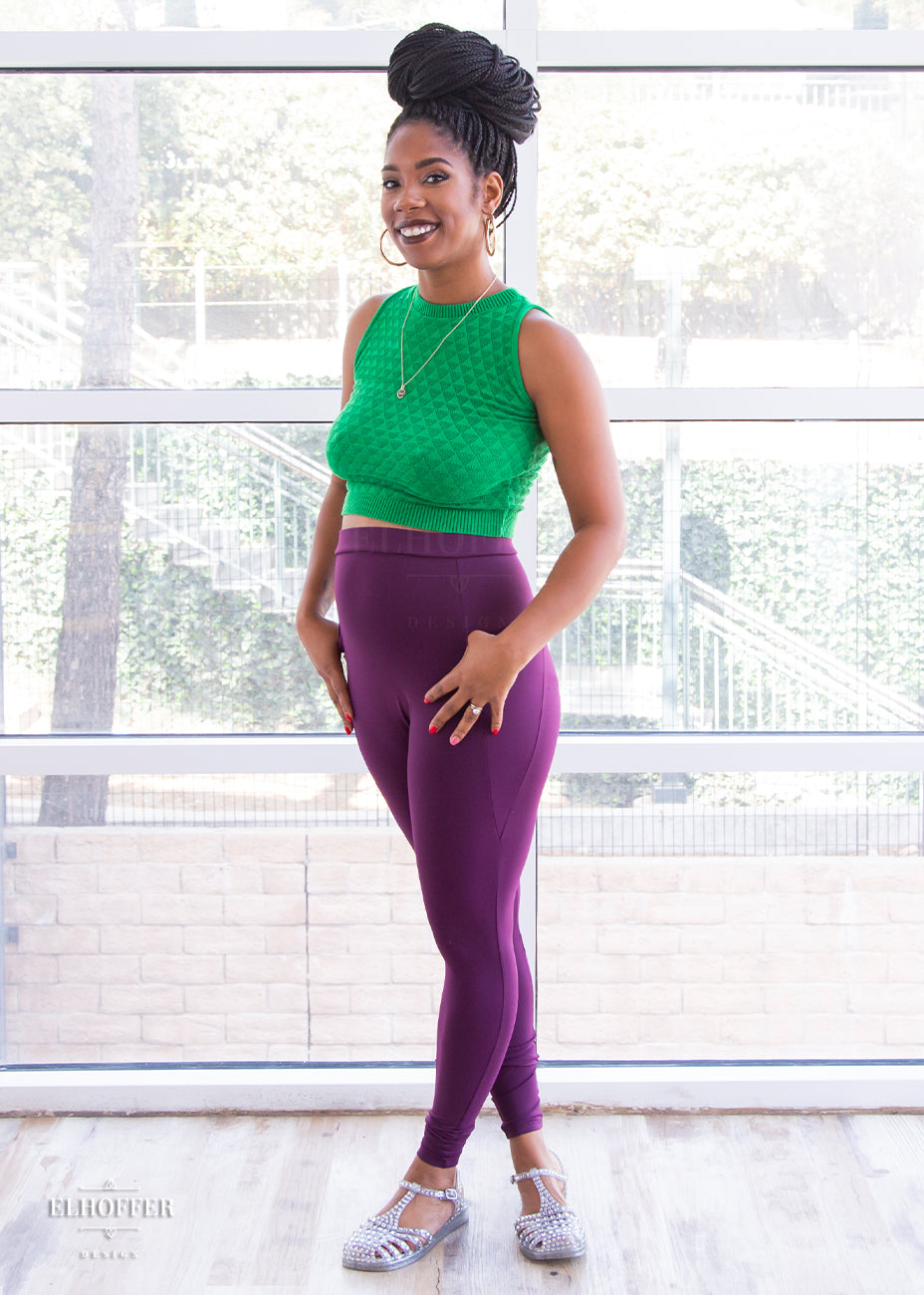 Krystina, a size small medium dark skinned model with long braids, is wearing a pair of high waisted leggings with a seamless front and side seam pockets in plum.