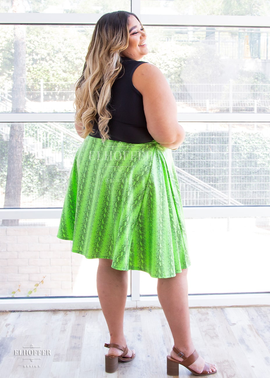 Cori, a medium skinned size 2XL model with brunette and blonde ombre hair, wears a high-waisted knee length full skirt with a fitted matching waistband encased with elastic in the croki green print. The croki green print is a lime green snake print.