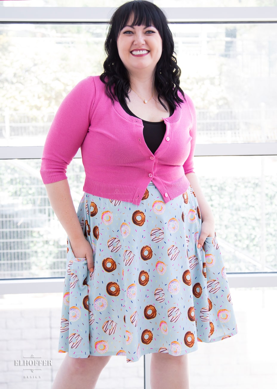 Bernadette, a fair skinned size large model with black hair and bangs, wears a high-waisted knee length full skirt with a fitted matching waistband encased with elastic in our donut stop believing print. The print has a light blue background and is covered in various donuts and sprinkles.