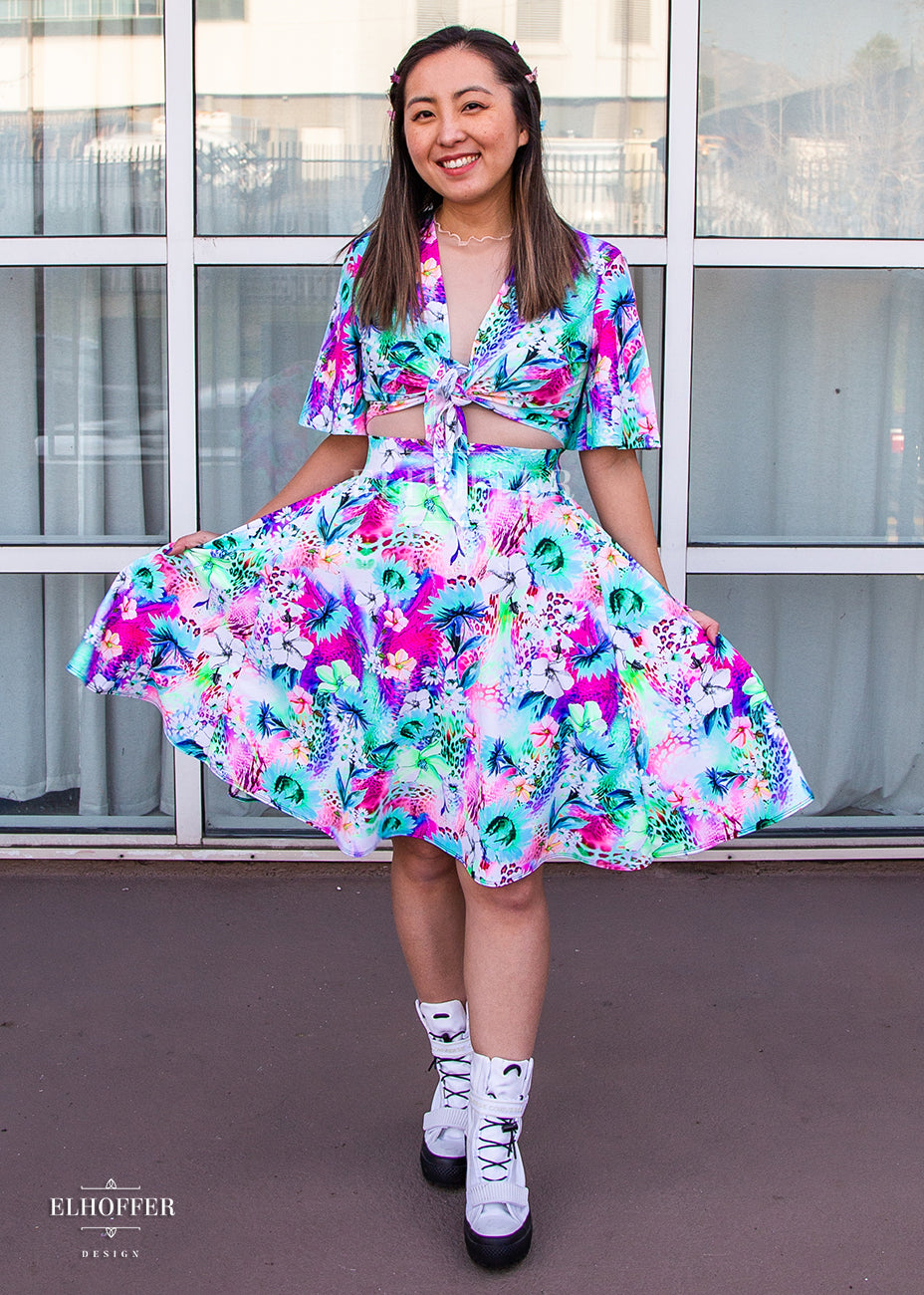 Alice, an olive skinned size XS model with highlighted brown hair, wears a high-waisted knee length full skirt with a fitted matching waistband encased with elastic in our rainbow jungle. The pattern is bright blue, pink and green with tropical flowers.