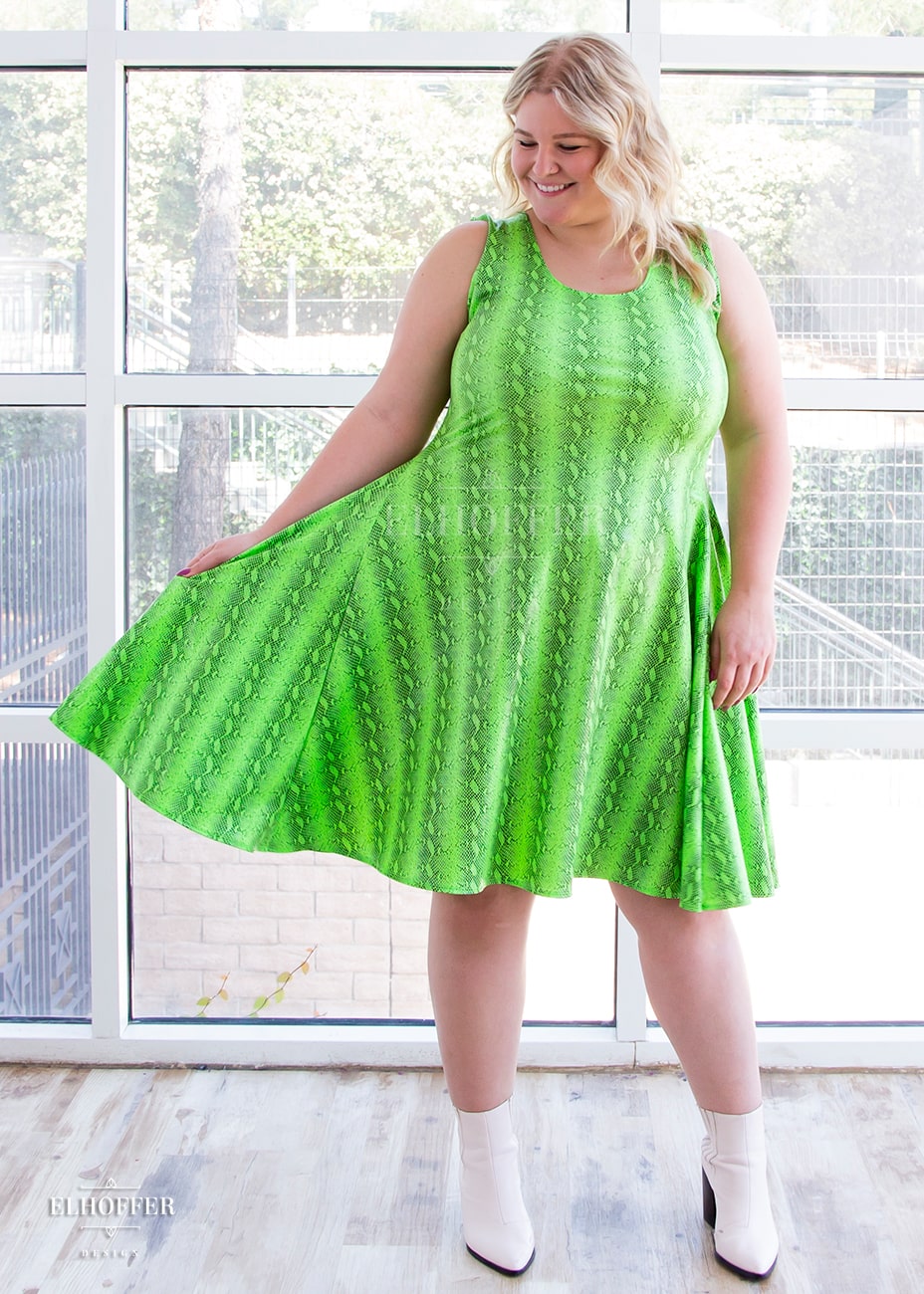 Sarah, a fair skinned size XL model with long blonde hair, wears a sleeveless knee length dress that is fitted in the torso and full in the skirt. The print is croki green, a lime green snake print. She holds out the side of her dress to show the fullness.