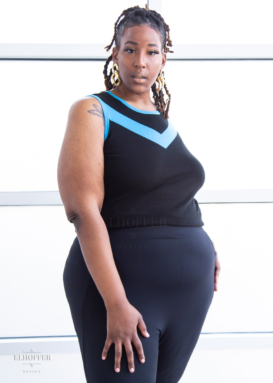 Myjah, a medium dark skinned 3xl model with shoulder length braids, is wearing a light weight sleeveless knit top.  The body of the top is mainly black with a bright teal chevron design across the chest and matching teal binding around the neckline and armholes.