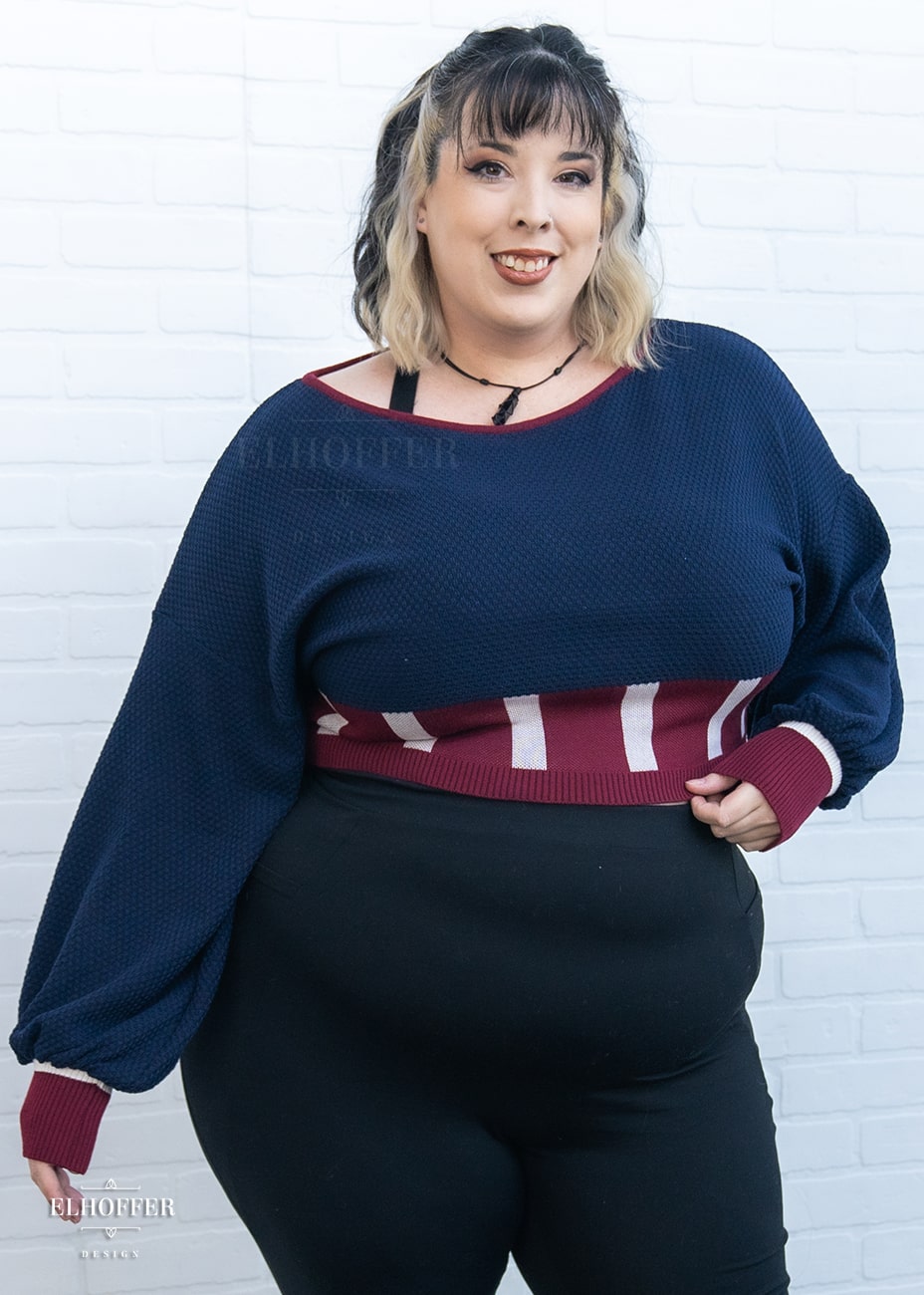 Katie Lynn, a size 2XL fair skinned model with black and blonde hair, is wearing the red, white and blue knit oversized crop top with bishop sleeves. The sleeves and top of the crop are blue with a waffle knit, the waist is vertical red and white stripes. The cuffs are mainly red with a white detail. There is also a red detail at the neckline.