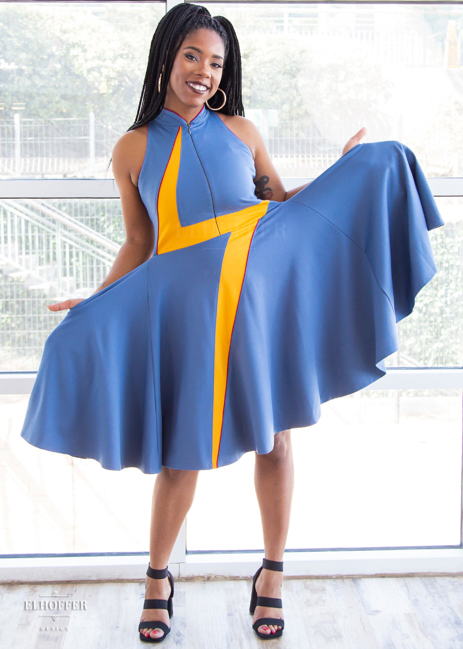 Krystina, a size small medium dark skinned model with long braids, holds out the full skirt of the high-necked sleeveless blue dress. The dress is piped in crimson and has a yellow lightening bolt emblazoned over the front of it. There is a zip down the middle down to the waist.