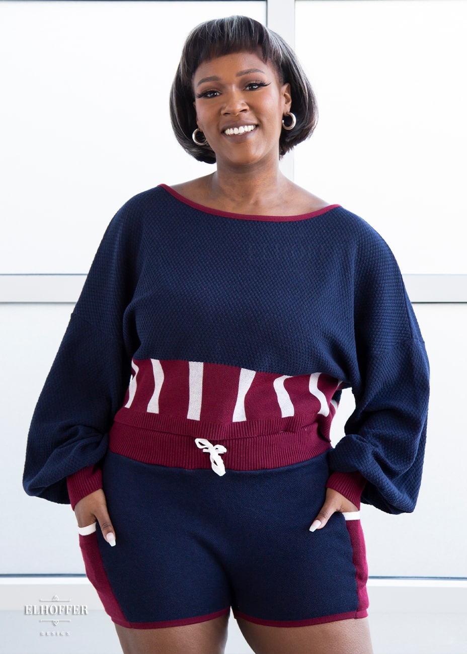 Lynsi, a medium dark skinned model with a dark brown and white bob, is wearing the red, white and blue knit oversized crop top with bishop sleeves. The sleeves and top of the crop are blue with a waffle knit, the waist is vertical red and white stripes. The cuffs are mainly red with a white detail. There is also a red detail at the neckline.