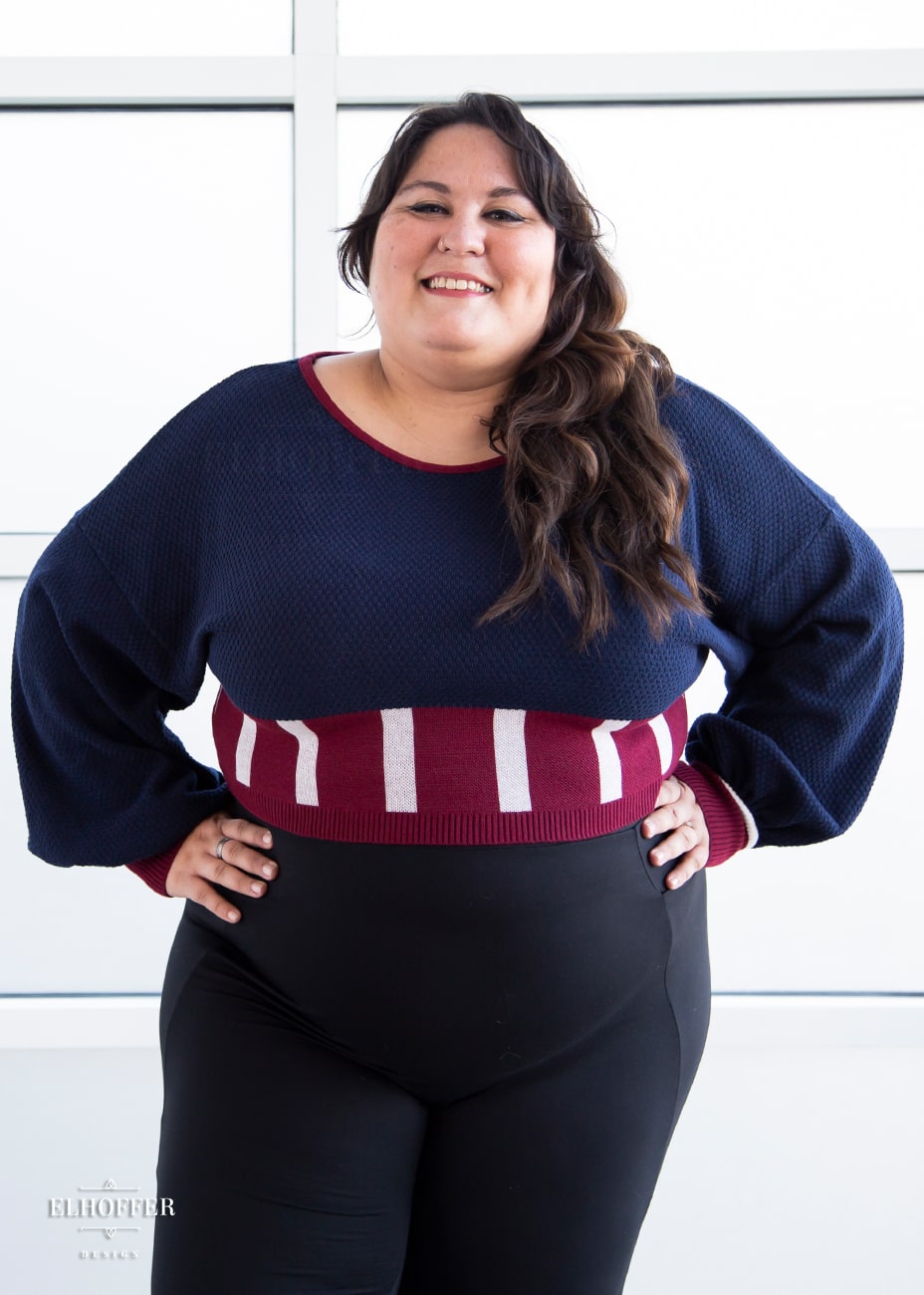 Alysia, a sun kissed skin size XL model with long brown hair, is wearing the red, white and blue knit oversized crop top with bishop sleeves. The sleeves and top of the crop are blue with a waffle knit, the waist is vertical red and white stripes. The cuffs are mainly red with a white detail. There is also a red detail at the neckline.