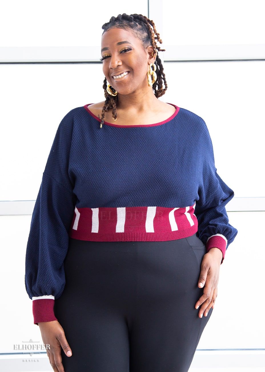 Myjah, a medium dark skinned 3xl model with shoulder length braids, is smiling while wearing the red, white and blue knit oversized crop top with bishop sleeves. The sleeves and top of the crop are blue with a waffle knit, the waist is vertical red and white stripes. The cuffs are mainly red with a white detail. There is also a red detail at the neckline.