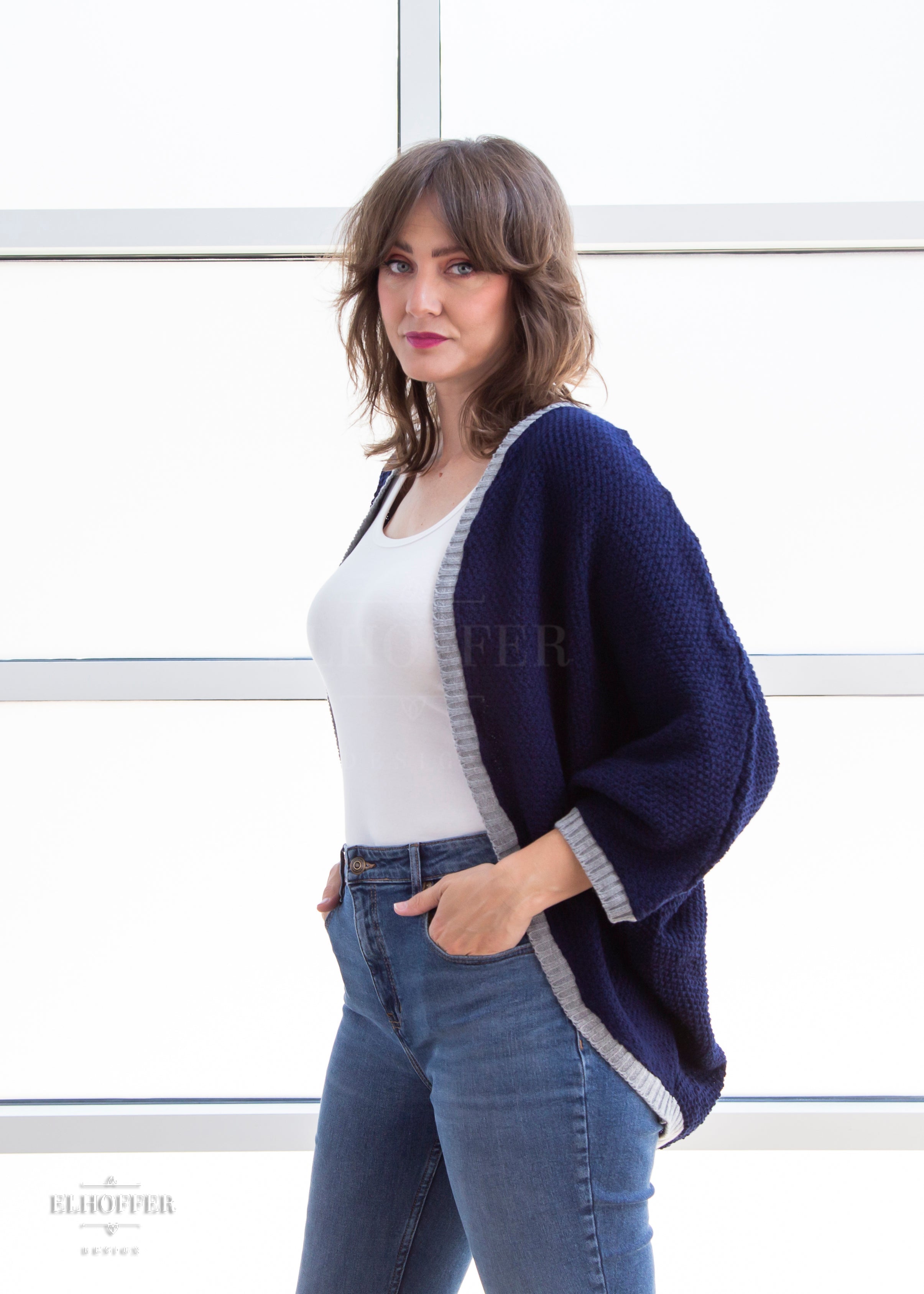 Ashley, a sun kissed skin M model with shoulder length feathered brown hair, is wearing a dark blue loose knit dolman with light grey ribbing around edges and cuffs. The dolman features 3/4 length sleeves and a magical script design (circle with T crossing through the circle) on the back.