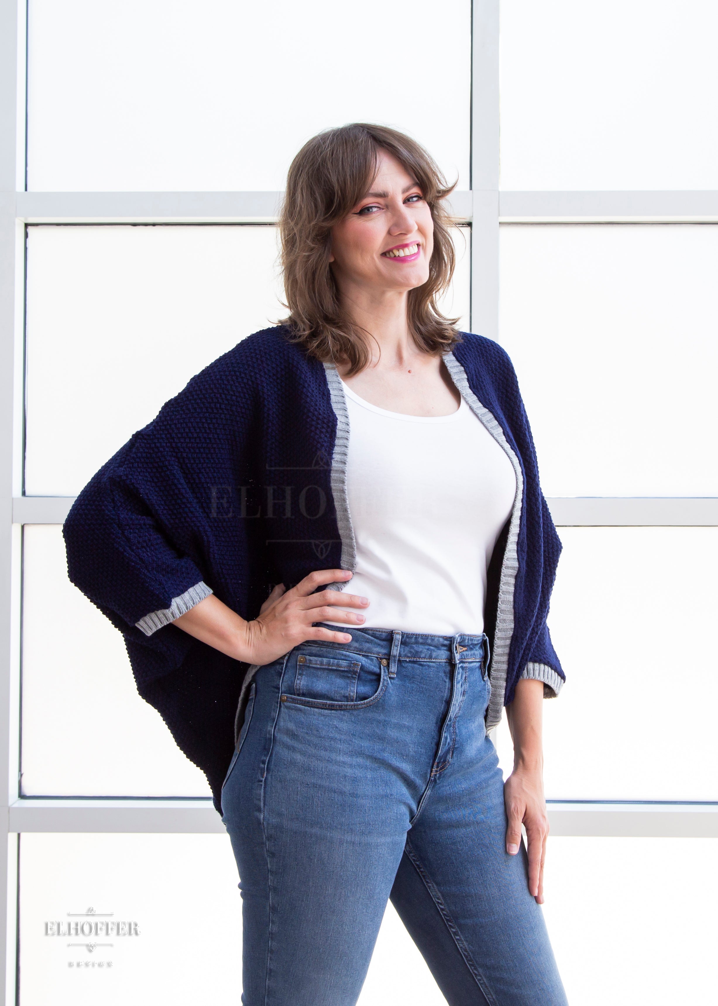 Ashley, a sun kissed skin M model with shoulder length feathered brown hair, is smiling while wearing a dark blue loose knit dolman with light grey ribbing around edges and cuffs. The dolman features 3/4 length sleeves and a magical script design (circle with T crossing through the circle) on the back.