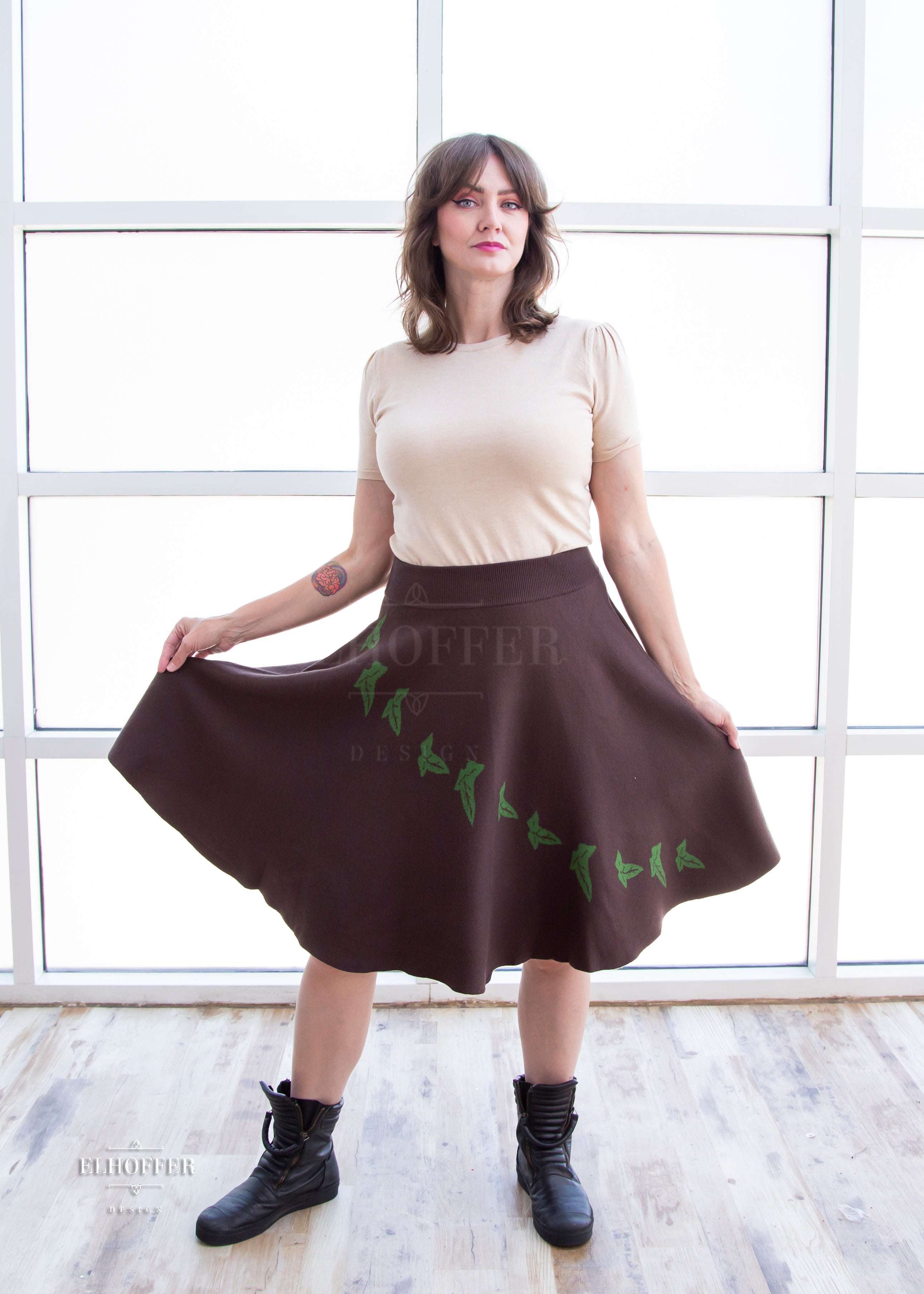 Ashley, a light skinned M/L model with brown hair, is wearing a brown knit skirt that hits just passed the knee and has a cascading green leaf design and an oatmeal colored lightweight knit t-shirt style top.