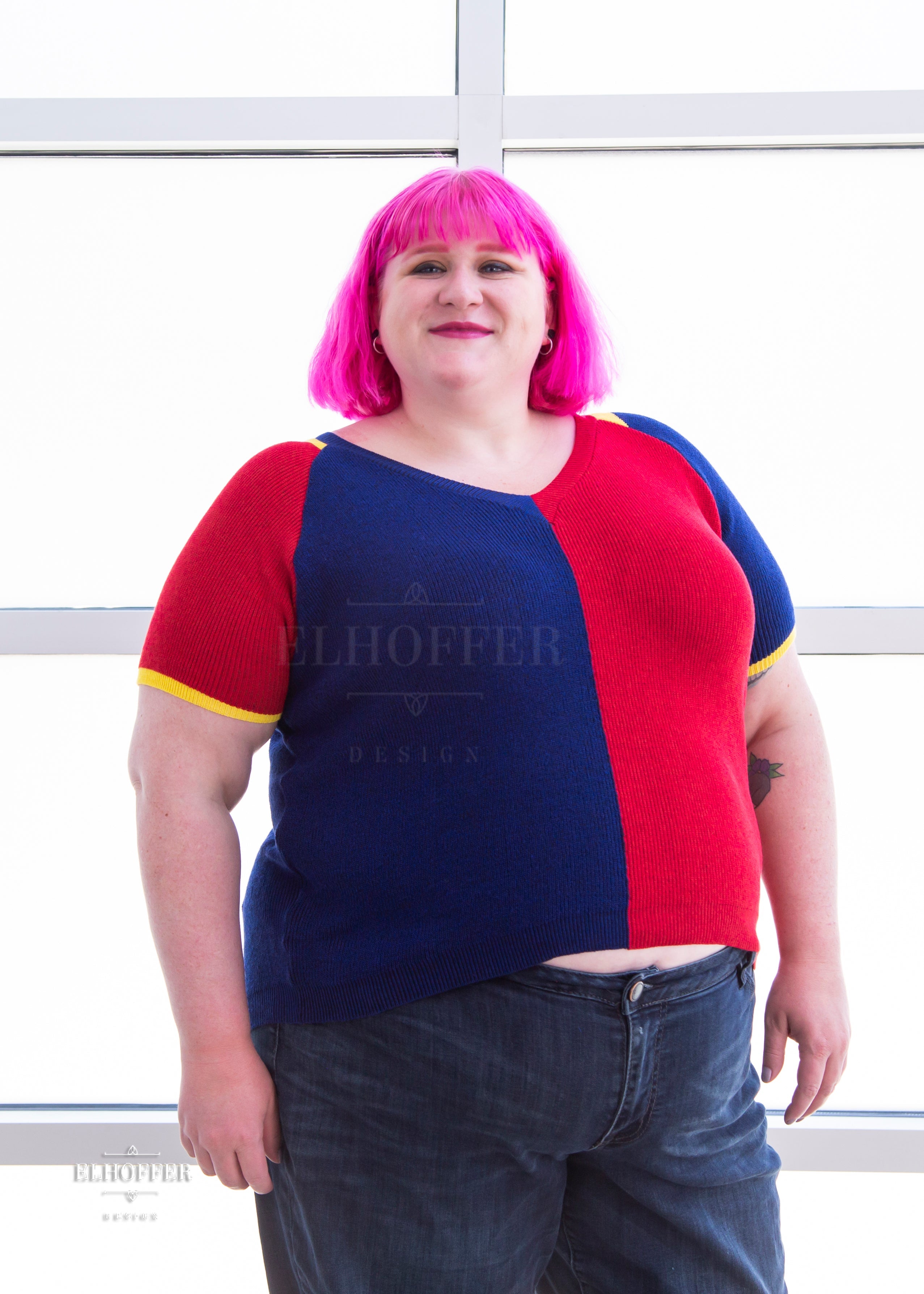 Logan, a fair skinned 3xl model with short pink hair with bangs, is wearing a short sleeve knit top with alternating blue and red colors. There is yellow detailing along the top of the shoulder and around the cuff of the sleeve.