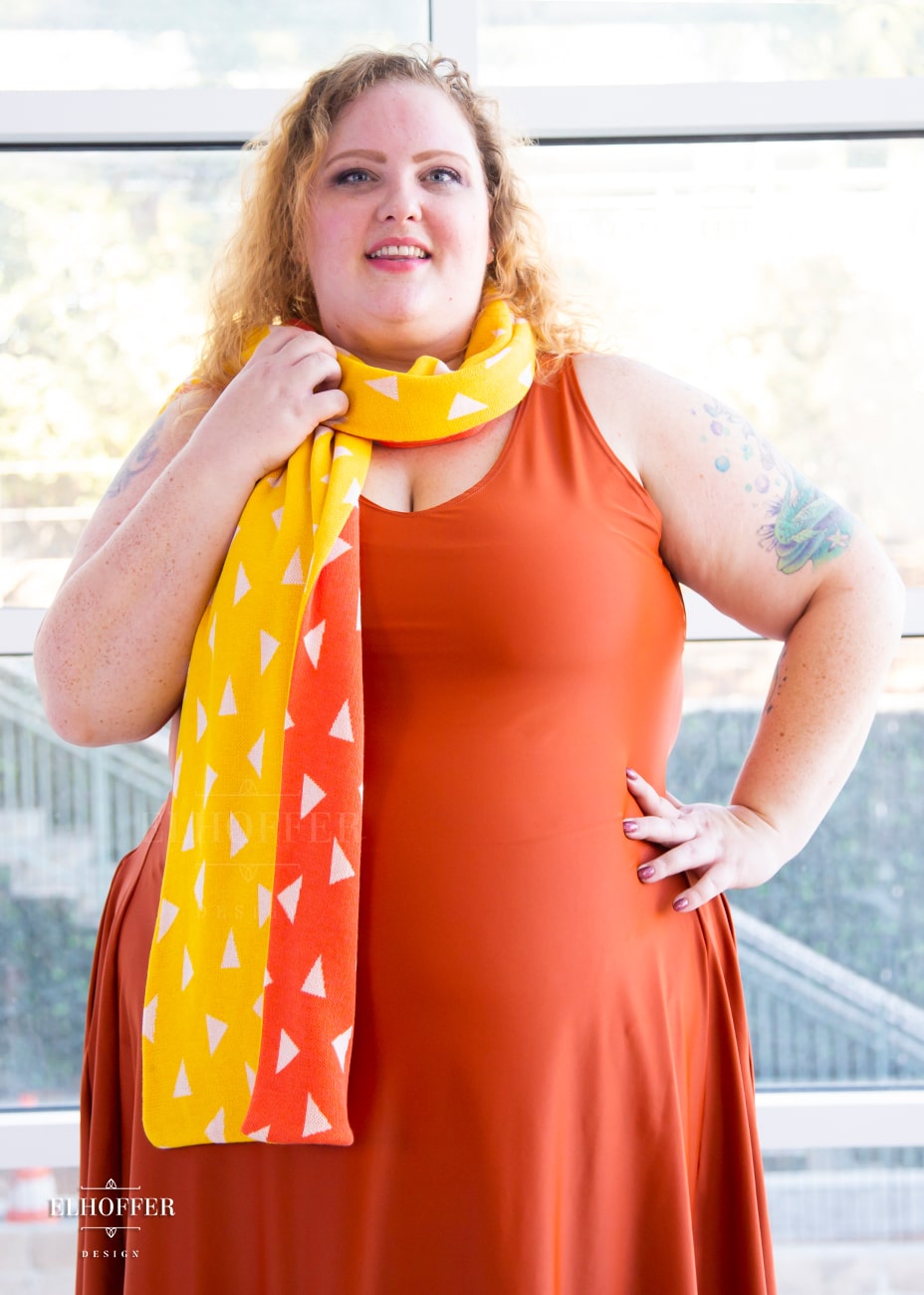 Bee, a fair skinned 3xl model with medium length curly blonde hair, is smiling while wearing a reversible knit scarf.  One side of the scarf is orange with white triangles and the other side is yellow with white triangles.