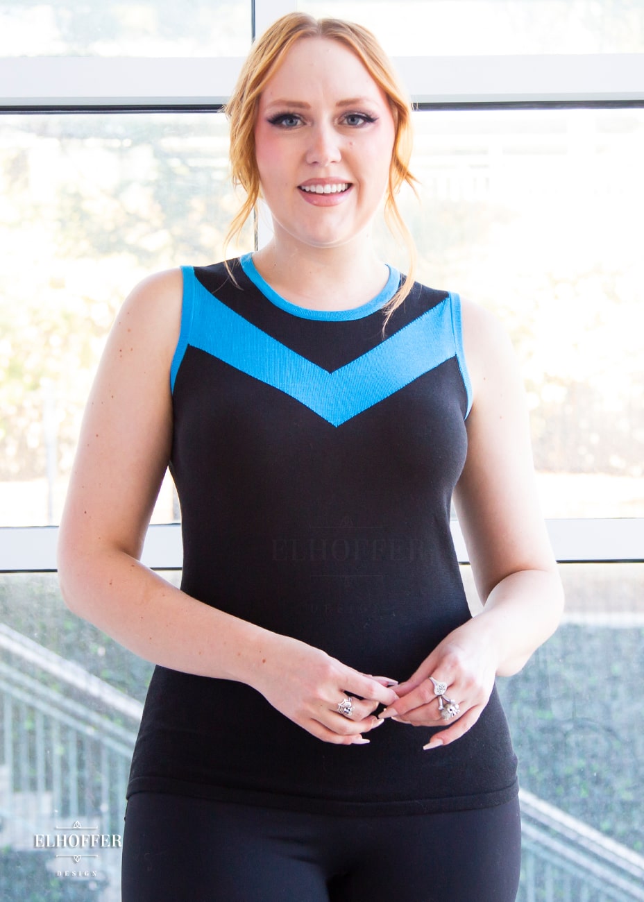 Kelsey, a fair skinned S model with strawberry blonde hair pulled back in a pony tail, is smiling while wearing a light weight sleeveless knit top. The body of the top is mainly black with a bright teal chevron design across the chest and matching teal binding around the neckline and armholes.