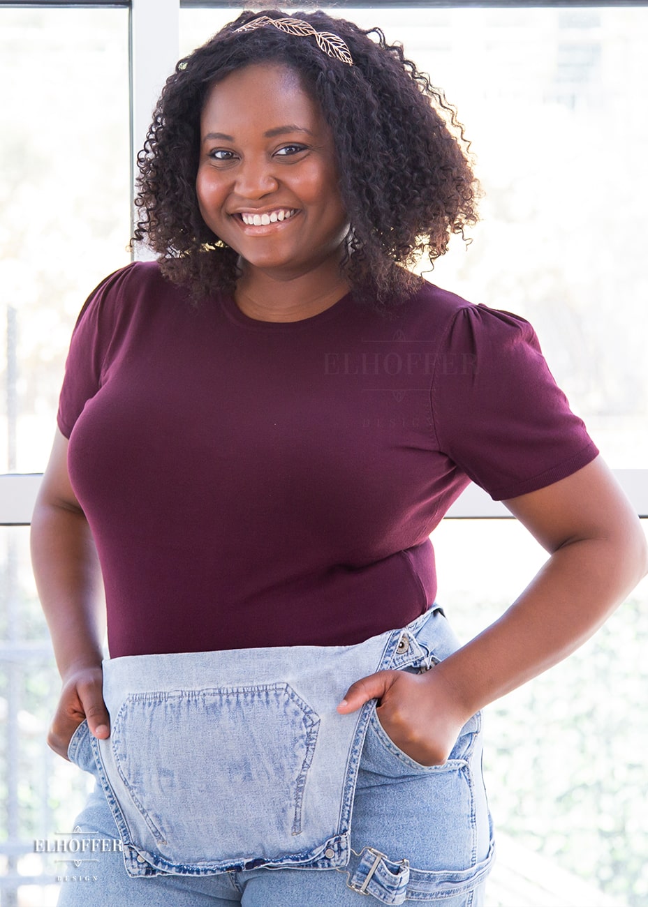 Maydelle, a medium dark skinned XL model with dark shoulder length tight curly hair, is smiling while wearing a short sleeve light weight dark reddish purple knit top. The top hits about mid hip in length and the sleeves have pleated gathering at the shoulders.