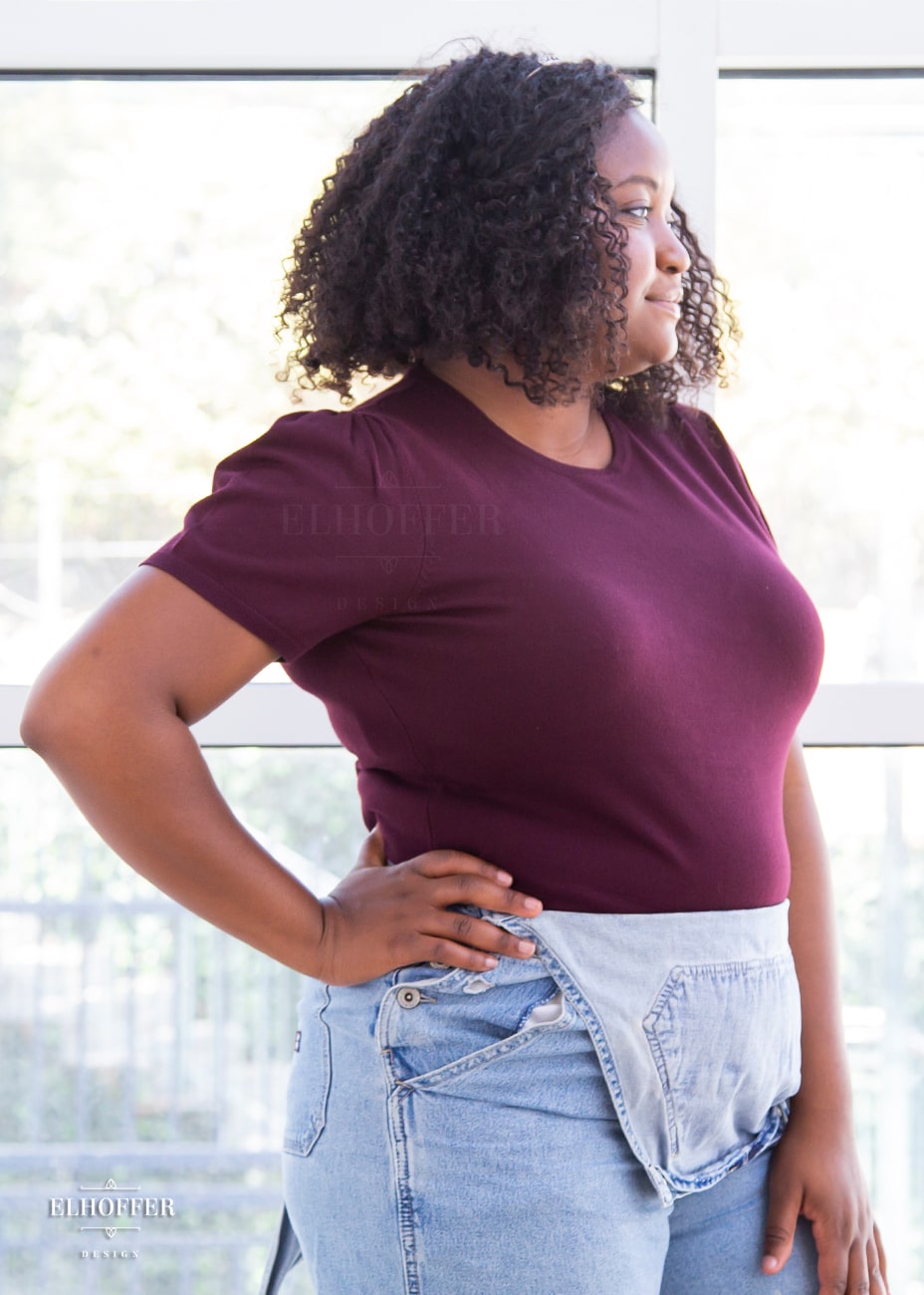 A side view of Maydelle, a medium dark skinned XL model with dark shoulder length tight curly hair, wearing a short sleeve light weight dark reddish purple knit top. The top hits about mid hip in length and the sleeves have pleated gathering at the shoulders.
