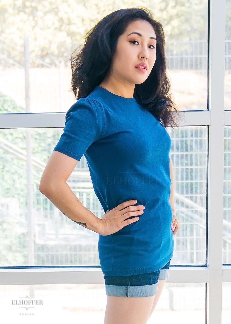 Kate, an olive skinned XS model with long dark hair, is wearing a blue knit short sleeve tee style top. The top has a high round neckline and the hem of the top falls to the hips. The shoulders are slightly gathered.