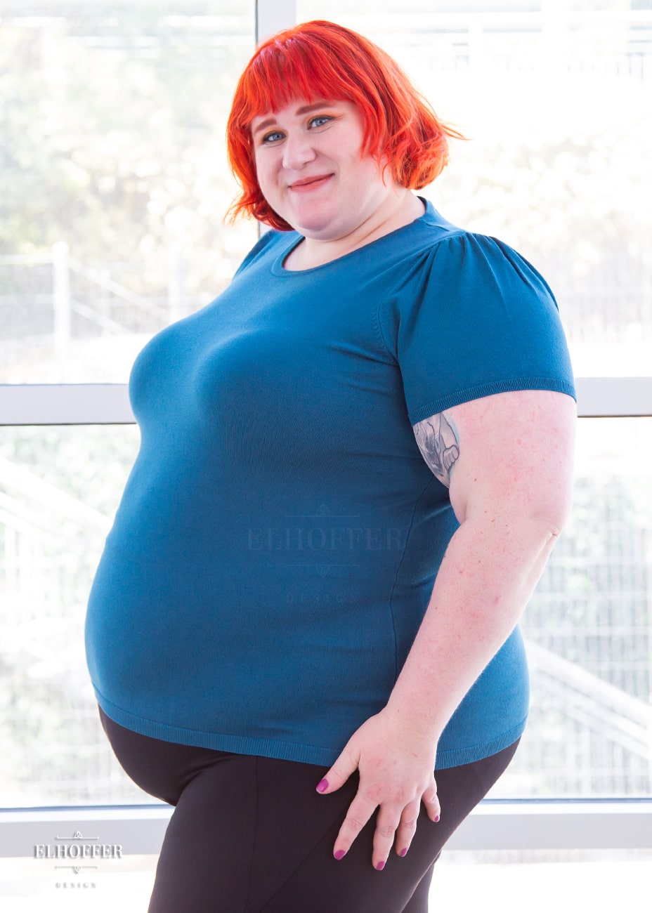 Logan, a fair skinned 3xl model with short orange hair with bangs, is wearing a short sleeve light weight peacock teal knit top. The top hits about mid hip in length and the sleeves have pleated gathering at the shoulders.