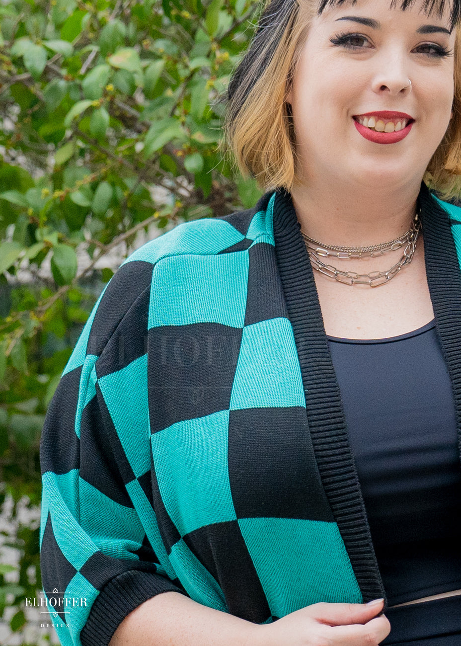 A close up of Katie Lynn, a fair skinned 2xl model with short black and blonde hair with bangs, to showcase the black and green chessboard pattern and black ribbing along front edge and cuffs.